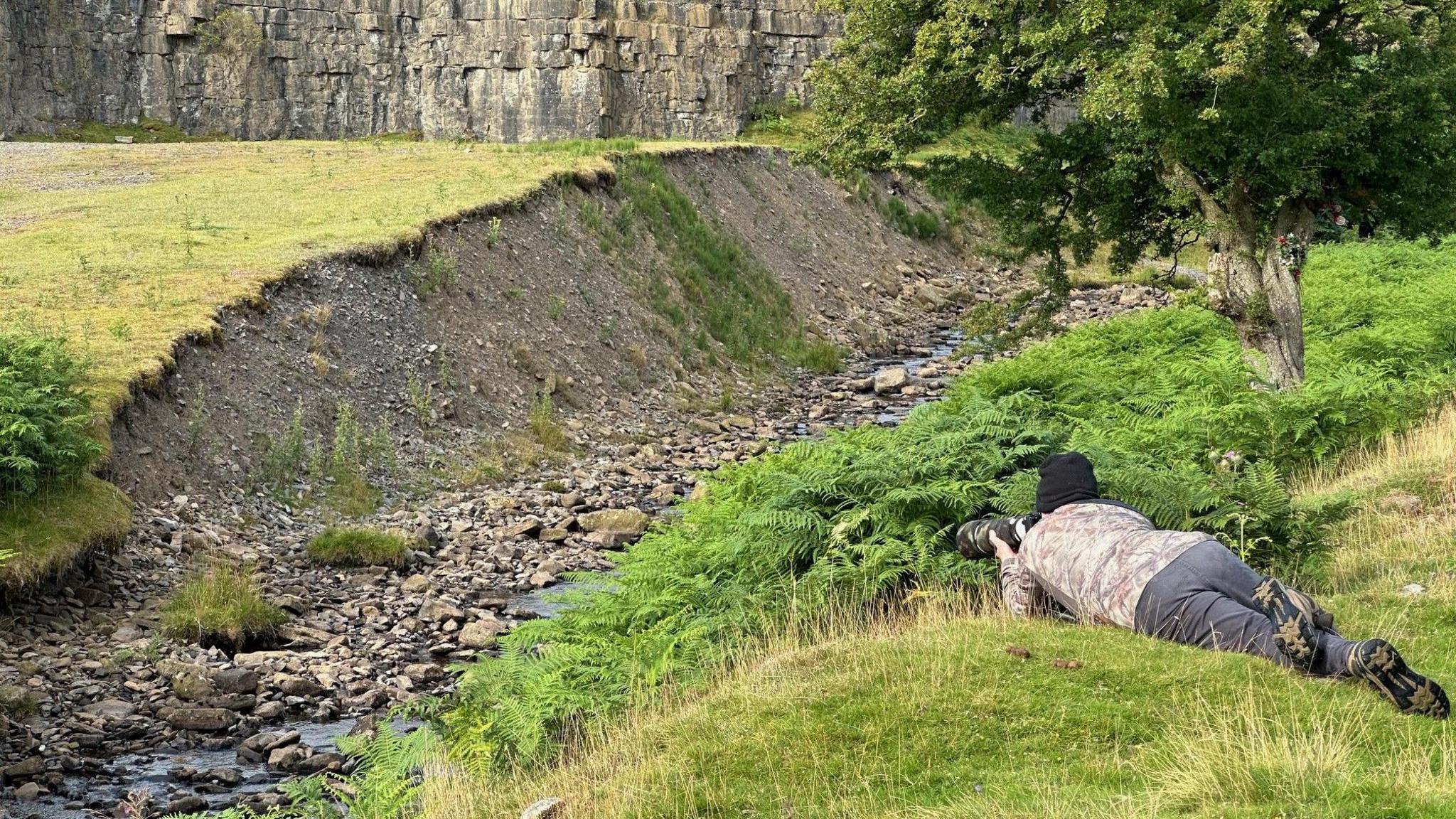 A man in a camouflage jacket lies on the bank of a stream pointing his camera to a small black and white bird which can just be made out in the bottom left of the picture