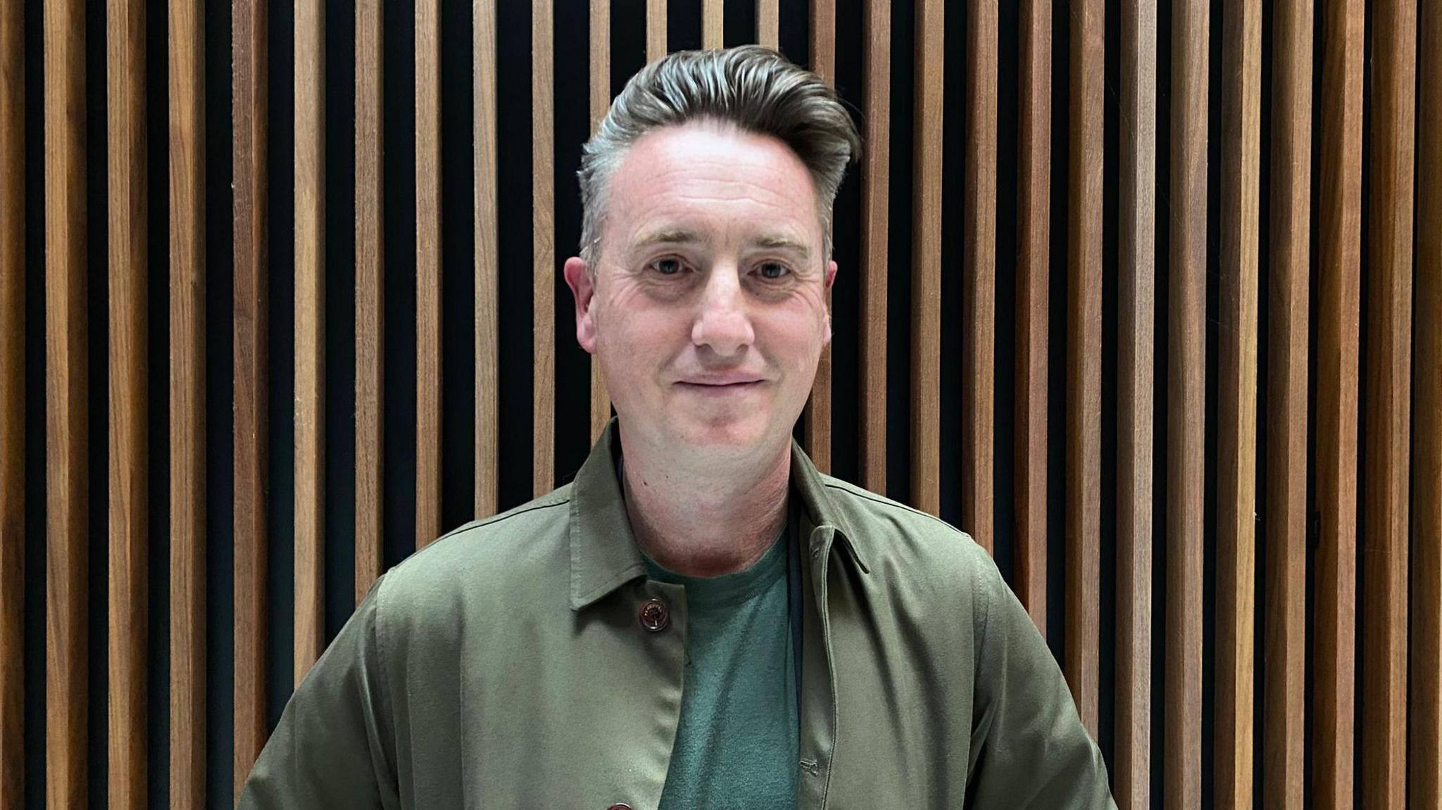 Dyn yn sefyll o flaen cefndir pren yn gwisgo crys T a chrys gwyrdd