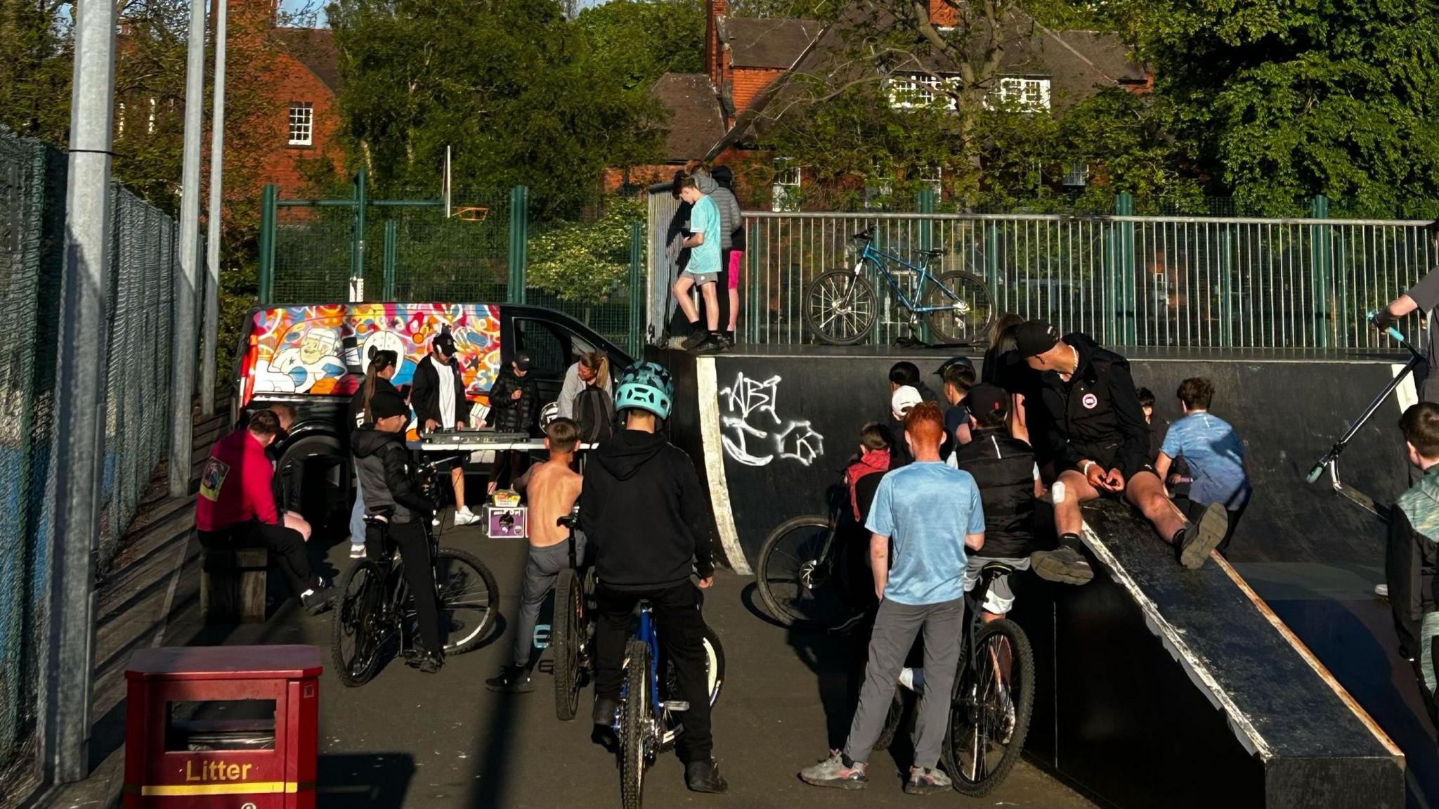 A previous park night. Dozens of young people are hanging out in a skate park. A lot of them have bikes. There is a colourful mural in the corner.
