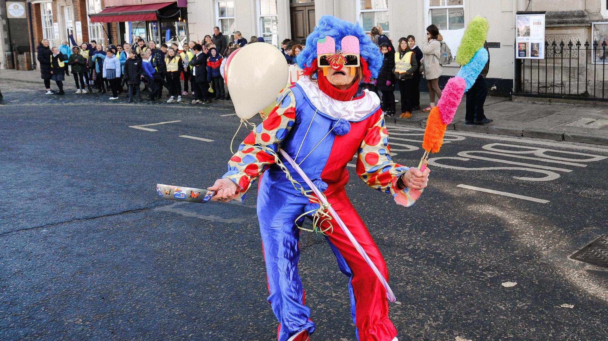 A person stands in the middle of the road in their fancy dress clown costume. It is a long-sleeved jumpsuit in metallic blue and red, with polka dot sleeves. They are holding multicoloured pom poms or dusters and balloons, with a silver frying pan which is covered in stickers. They have a bright blue wig on and large sunglasses in the shape of pink ice lollies.