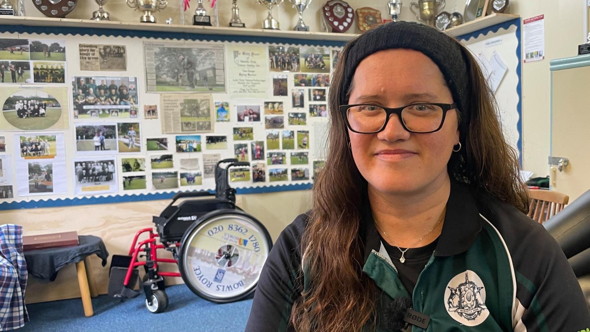 A woman with long brown hair and a black headband wearing black glasses and a green jersey. She is sitting in a club room surrounded by trophies and photographs.