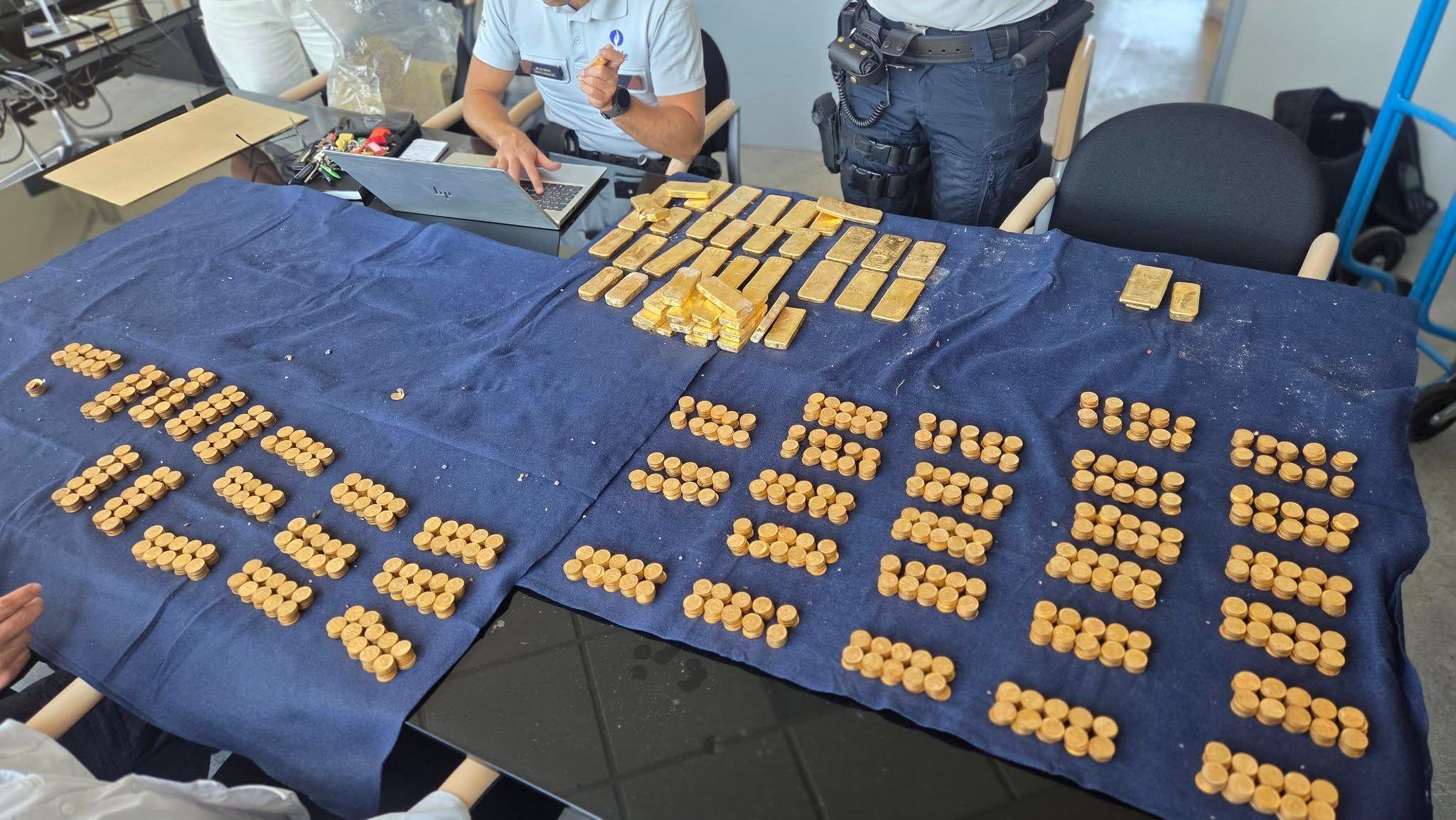 Hundreds of gold coins and tens of gold bars laid out in stacks on a table covered with dark blue fabric. Two police officers can be seen sat at the table, with one on his laptop.