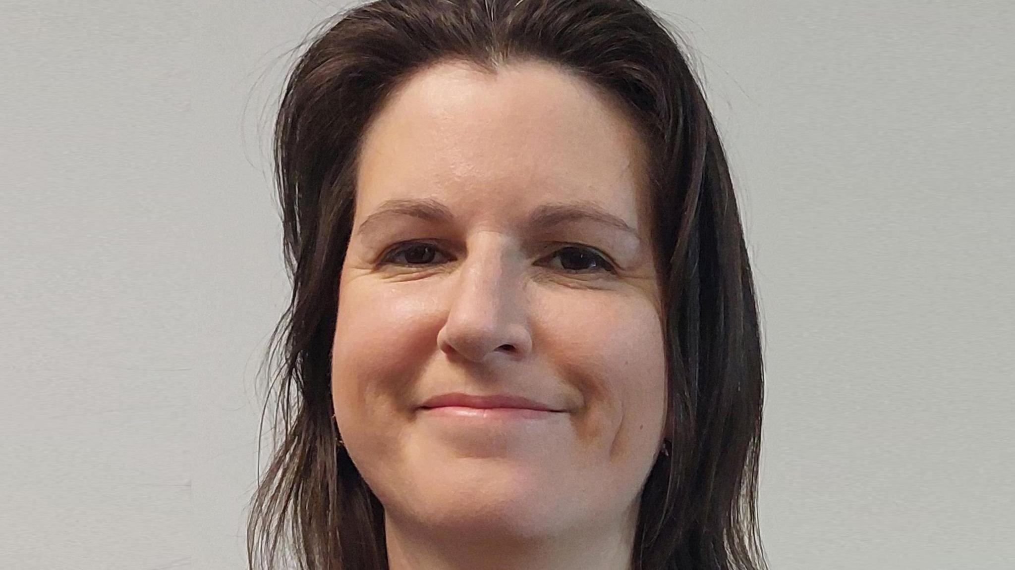 Woman with dark hair grinning for an official portrait photograph in front of a grey background.