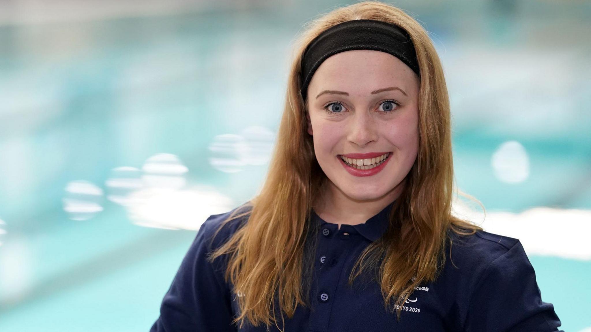 Ellie Robinson standing in front of a pool and smiling. She has brown hair and wearing a headband