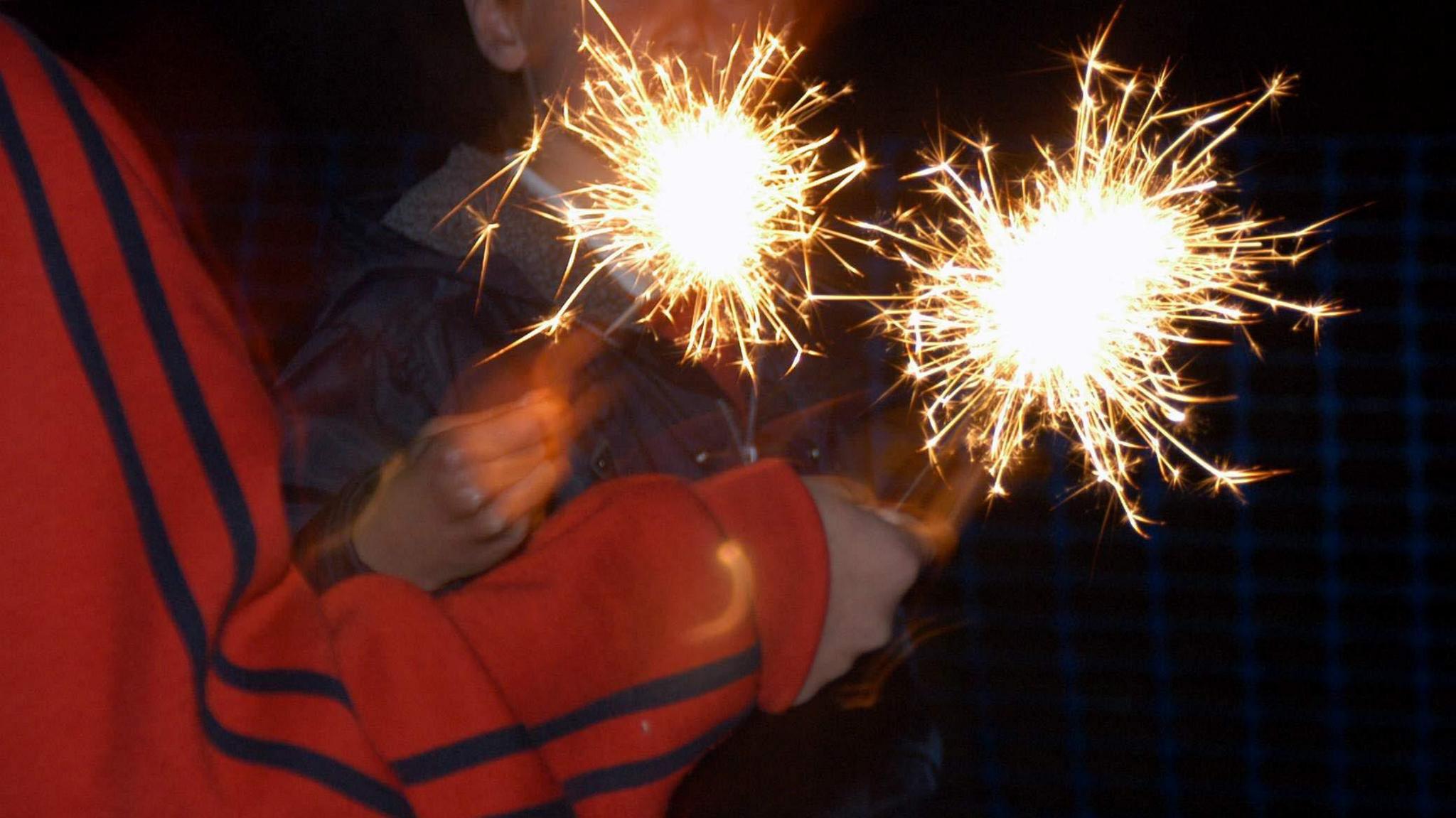A close up of two sparklers, one person with a red hoodie holds one of them it is dark outside.