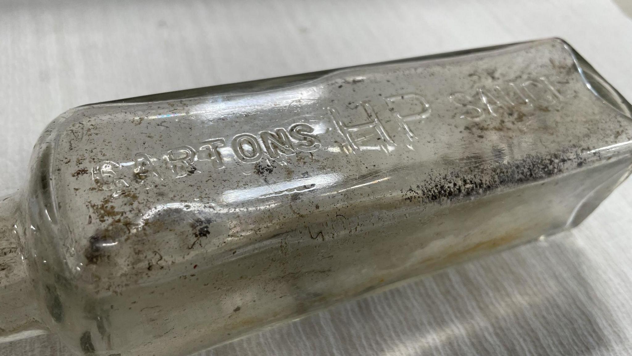 A glass bottle, marked Gartons HP Sauce. It is clearly very old and is covered in a layer of dirt.