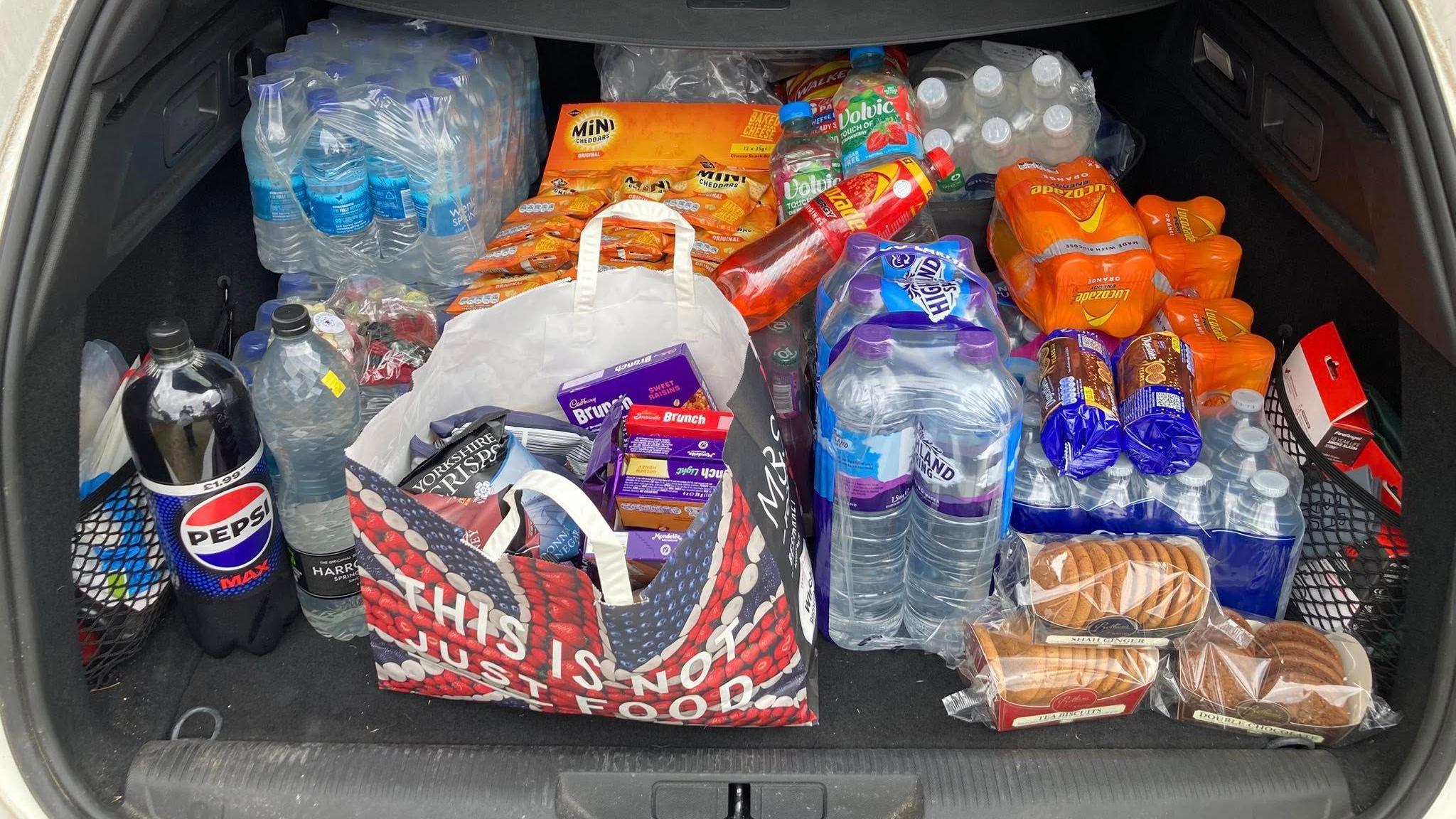 The open boot of a car filled with dozens of drinks bottles, packets of biscuits, chocolate bars and crisps.