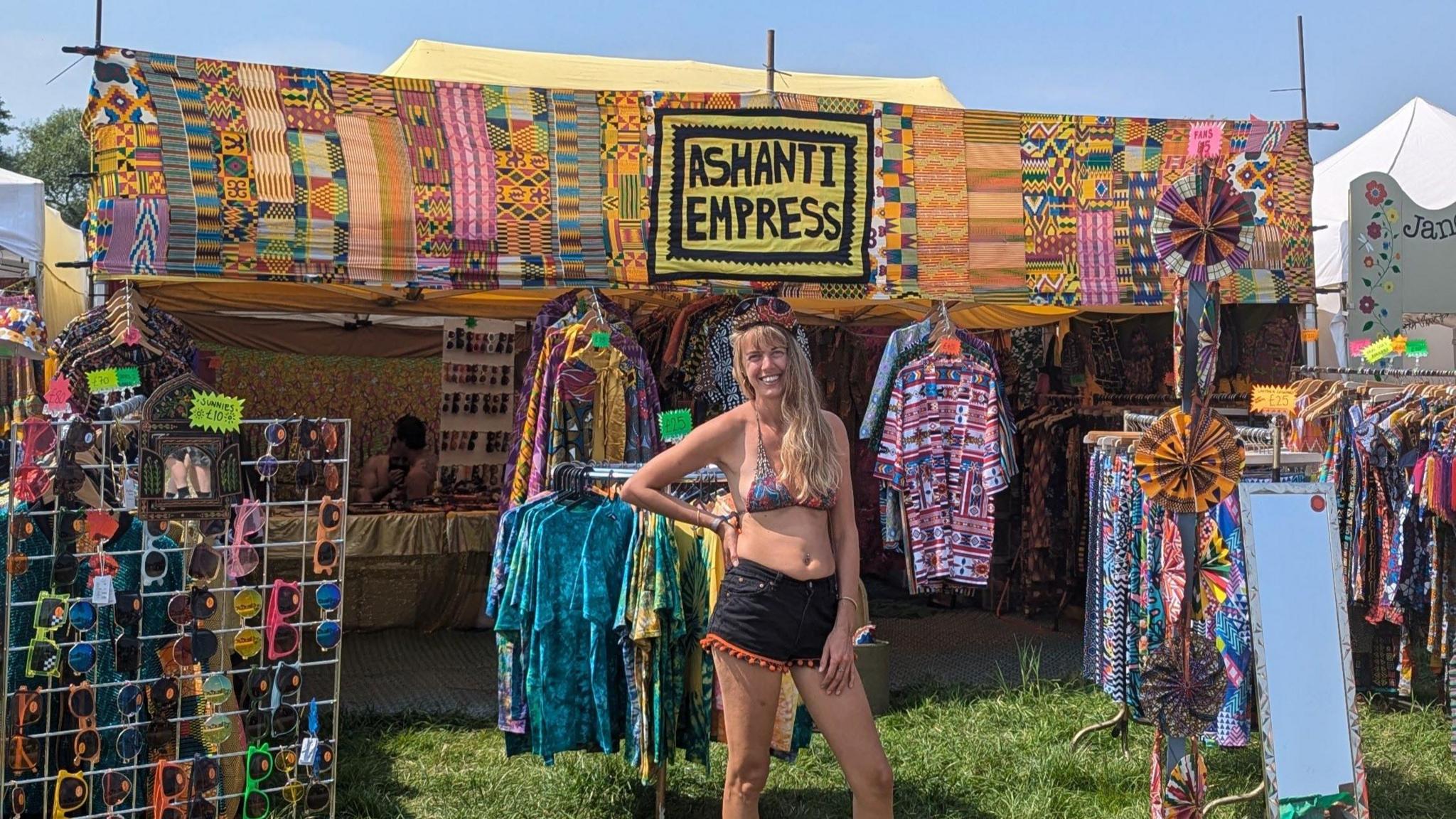 Ruby Maya poses for a picture in front of her colourful stall, with rails of clothing and a stand covered in sunglasses. A banner with the words 'Ashanti Empress' sewn into it runs along the top of the stall