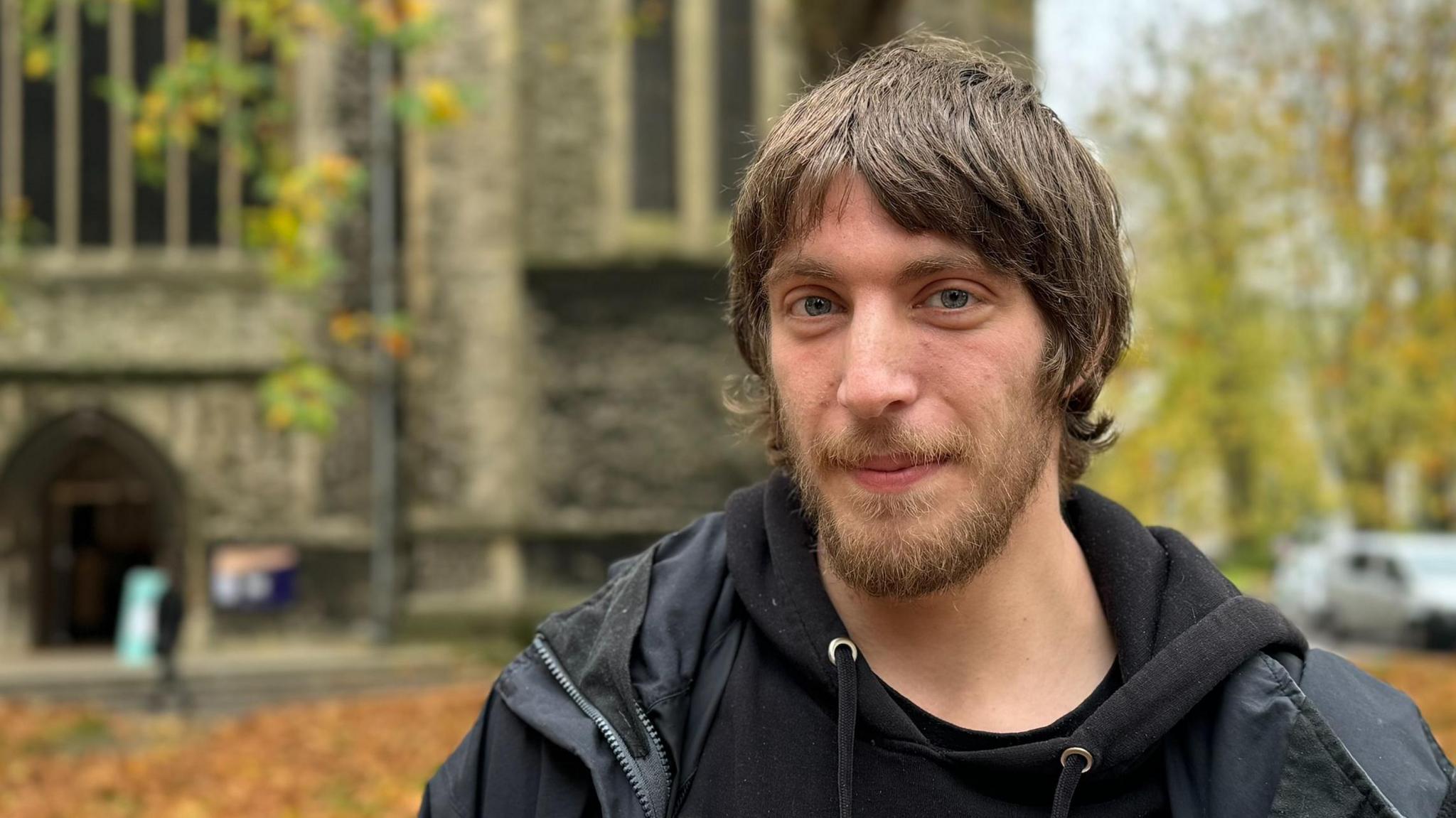 Ryan stands in front of St Mary's Church in Southampton, which is blurred in the background. He has medium length brown hair and a beard. He wears a black hoodie and a black raincoat over the top.