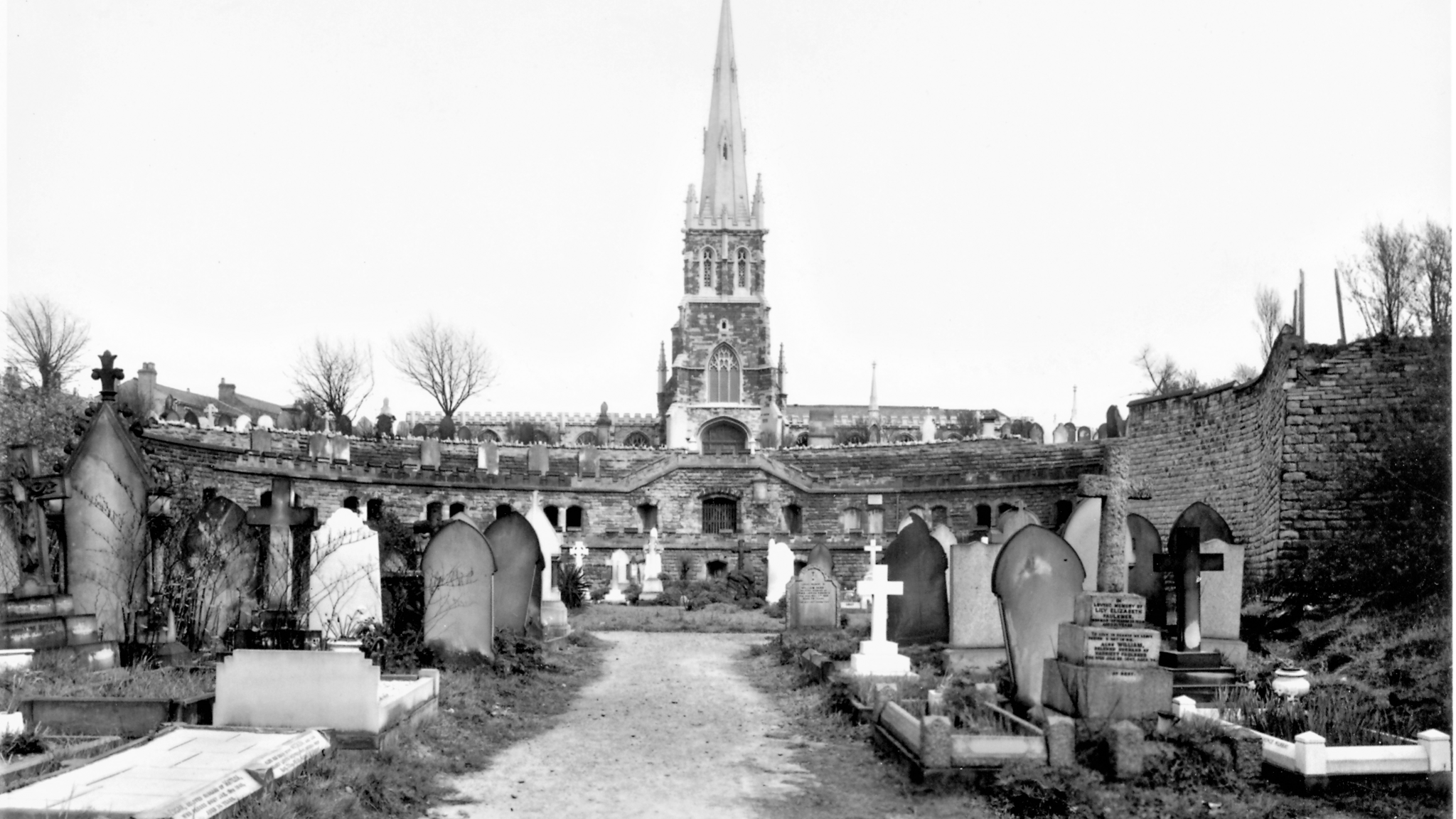 A black and white photograph showing a church spire in the centre of the image surrounded by grave stones of different shapes and sizes. Below the church are two levels with openings to the catacombs. 