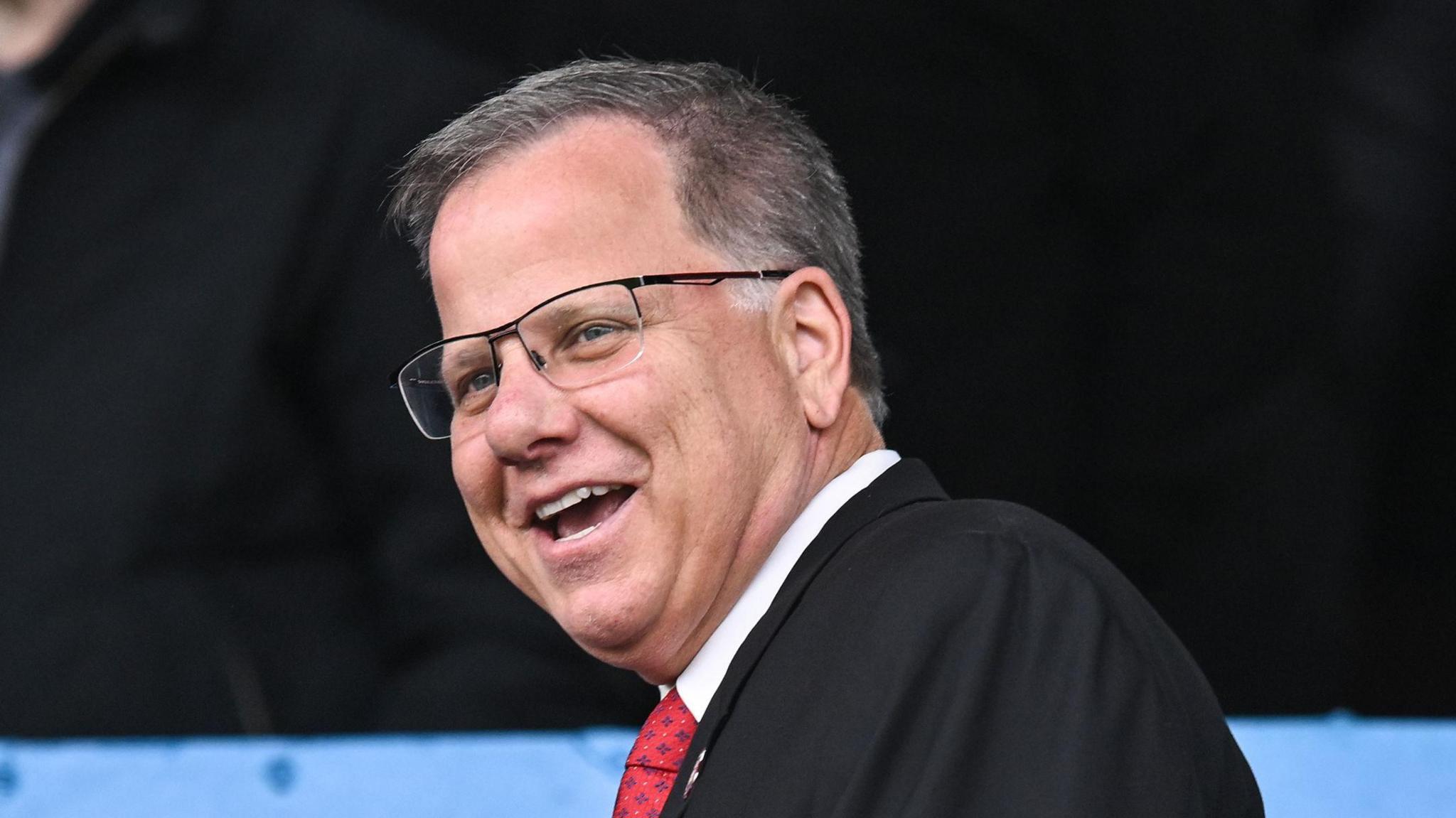 Tom Piatak, wearing a suit with a patterned red tie and glasses, about to take a seat at Carlisle's Brunton Park 