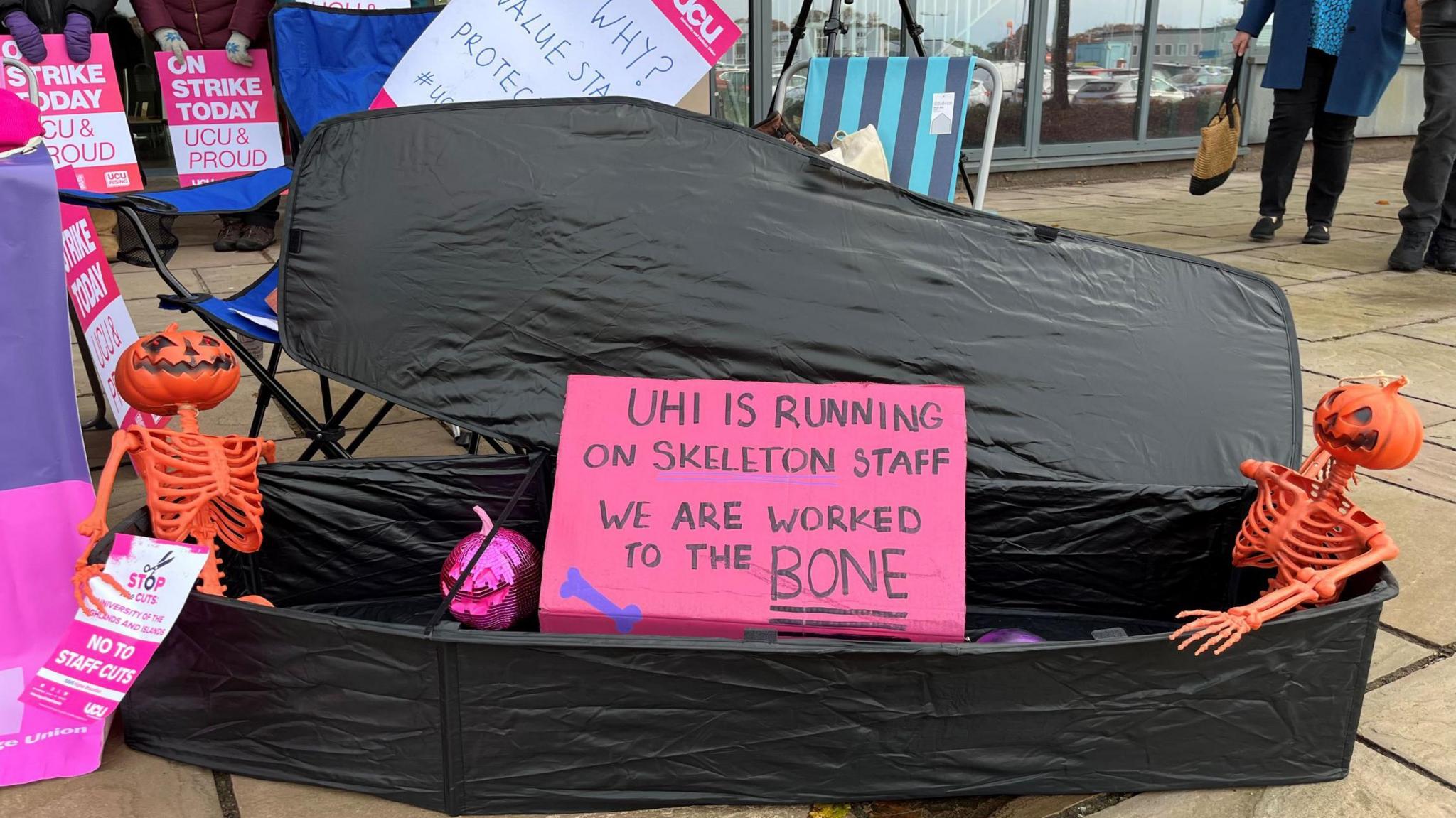 A mock coffin has been placed in front of the UHI Executive Office. There are two orange skeletons in it with pumpkin heads. A sign reads "the UHI is running on skeleton staff. We are being worked to the bone."