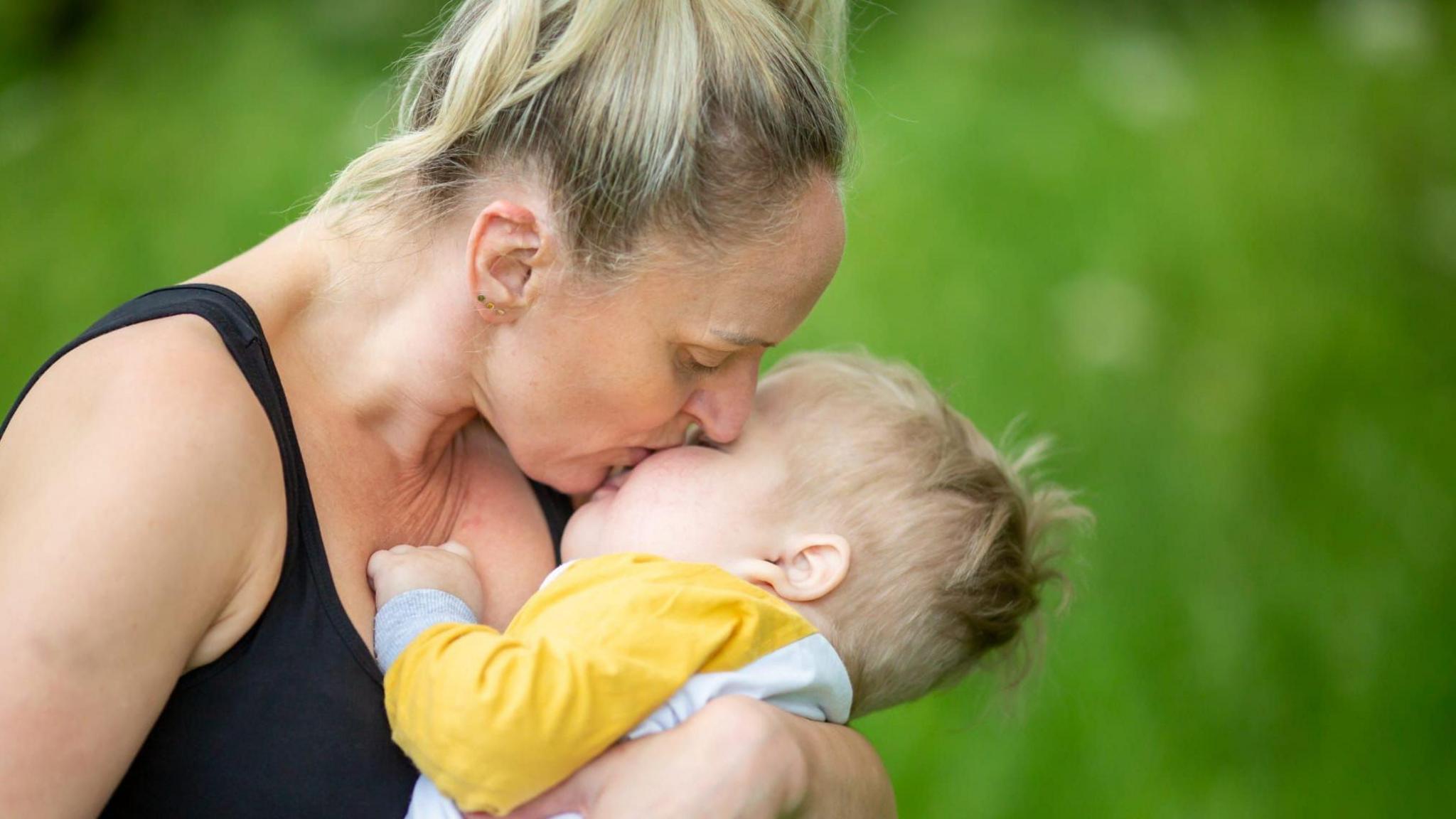 An image of Ezra being held by his mother as she kisses him on the cheek. Suzanne, his mother, is where a black tank top and her blonde hair is up in a ponytail. She has gold stud earrings. Ezra also has blonde hair and is wearing a white long-sleeve top with yellow sleeves. The camera is focused on them, but you can see grass in the background.
