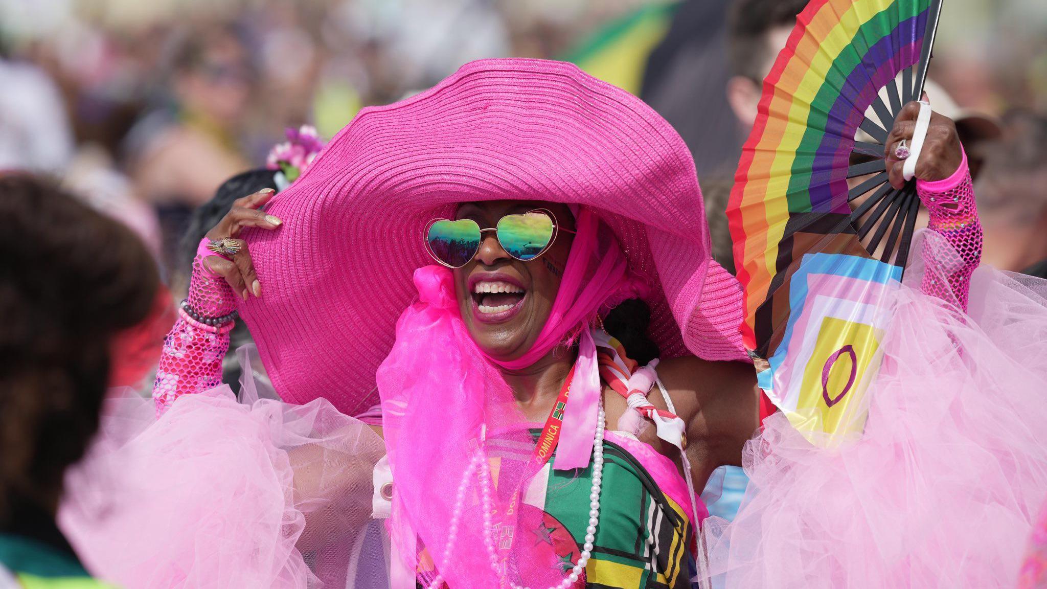 A person dressed all in pink. They have a large hat on and are holding a fan with the trans and pride flags on. They are wearing heart-shaped sunglasses.