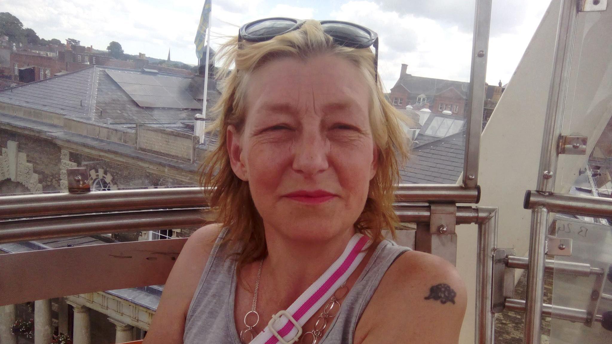 Dawn Sturgess, pictured smiling at the camera from what appears to be a viewing platform on the top of a building. She has short blonde/auburn hair, pushed back from her face by glasses, and wears a grey tank top and a pink and white bag strap across her torso. She has a small tattoo on her shoulder. 