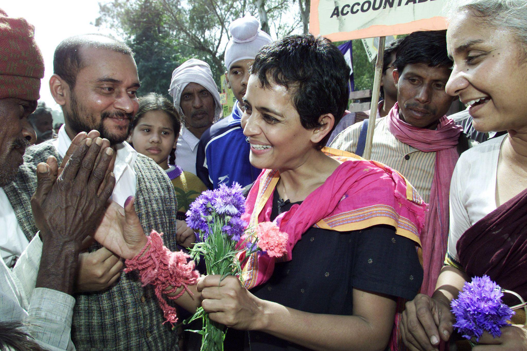 Arundhati Roy smiling and waving at a man as she holds a bunch of flowers in her right hand
