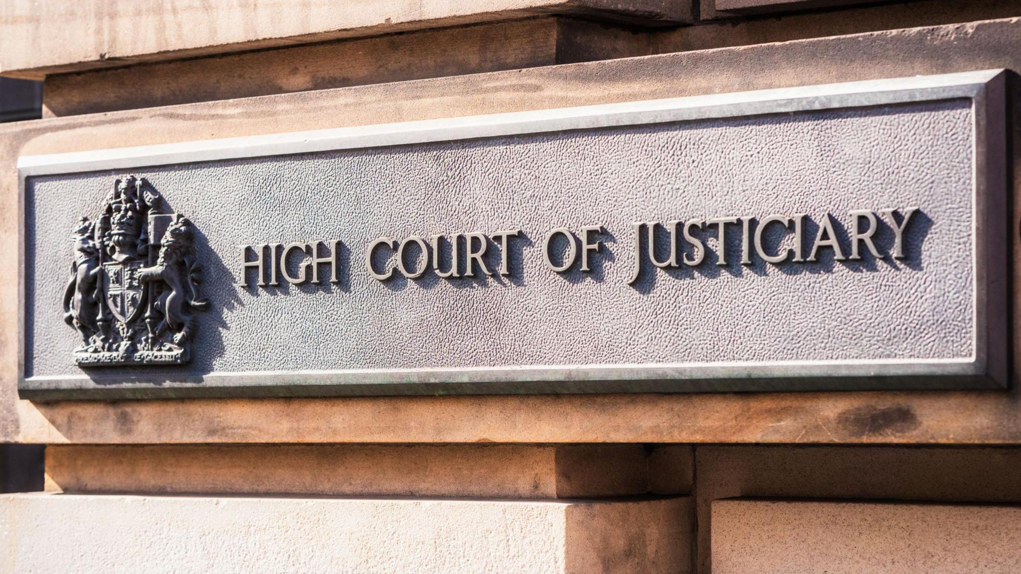 A rectangle-shaped grey sign on an exterior, stone block wall of the High Court in Edinburgh. The sign has a royal crest and the words: "High Court of Justiciary".