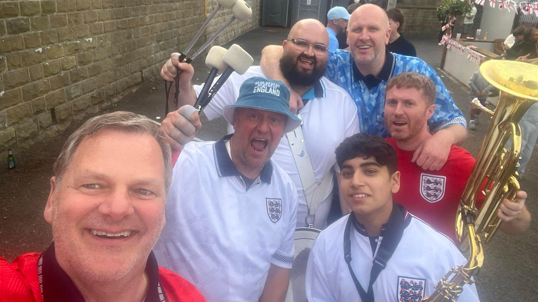 John Hemmingham, who has blue eyes and grey hair, pictured with some members of the England band. The other members are holding instruments including a saxophone, a drum and a trombone.