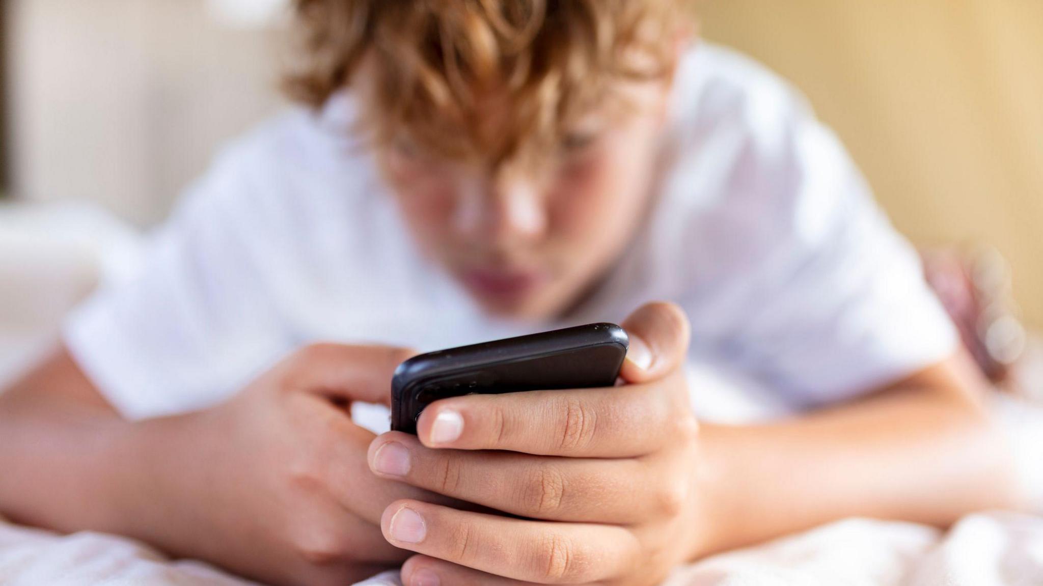 A young boy uses a smartphone. He wears a white t-shirt and lies on a bed. 