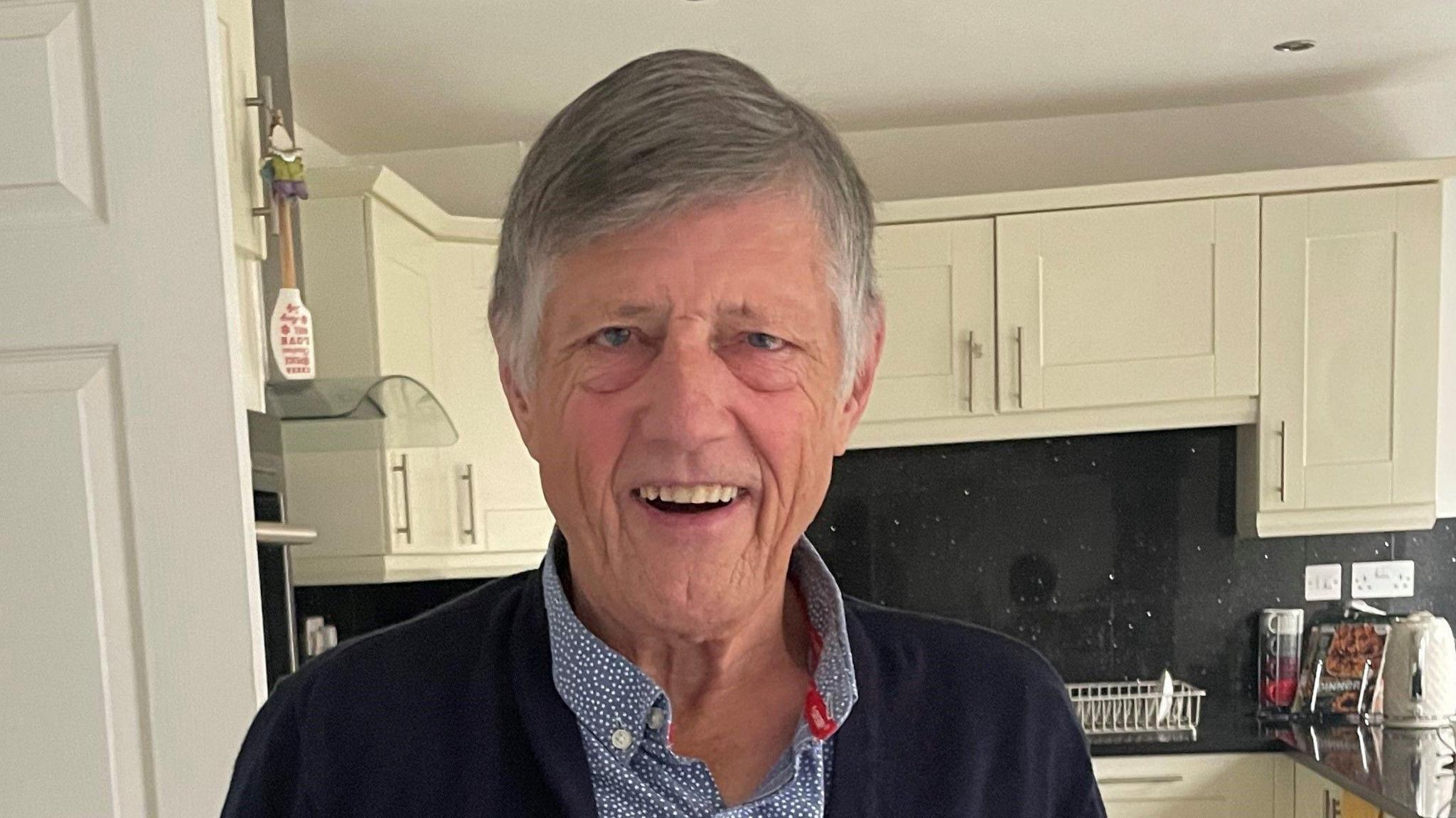 John Hardy smiles at the camera. He is clean shaven with grey hair and wearing a smart blue shirt, and is standing in a cream coloured kitchen