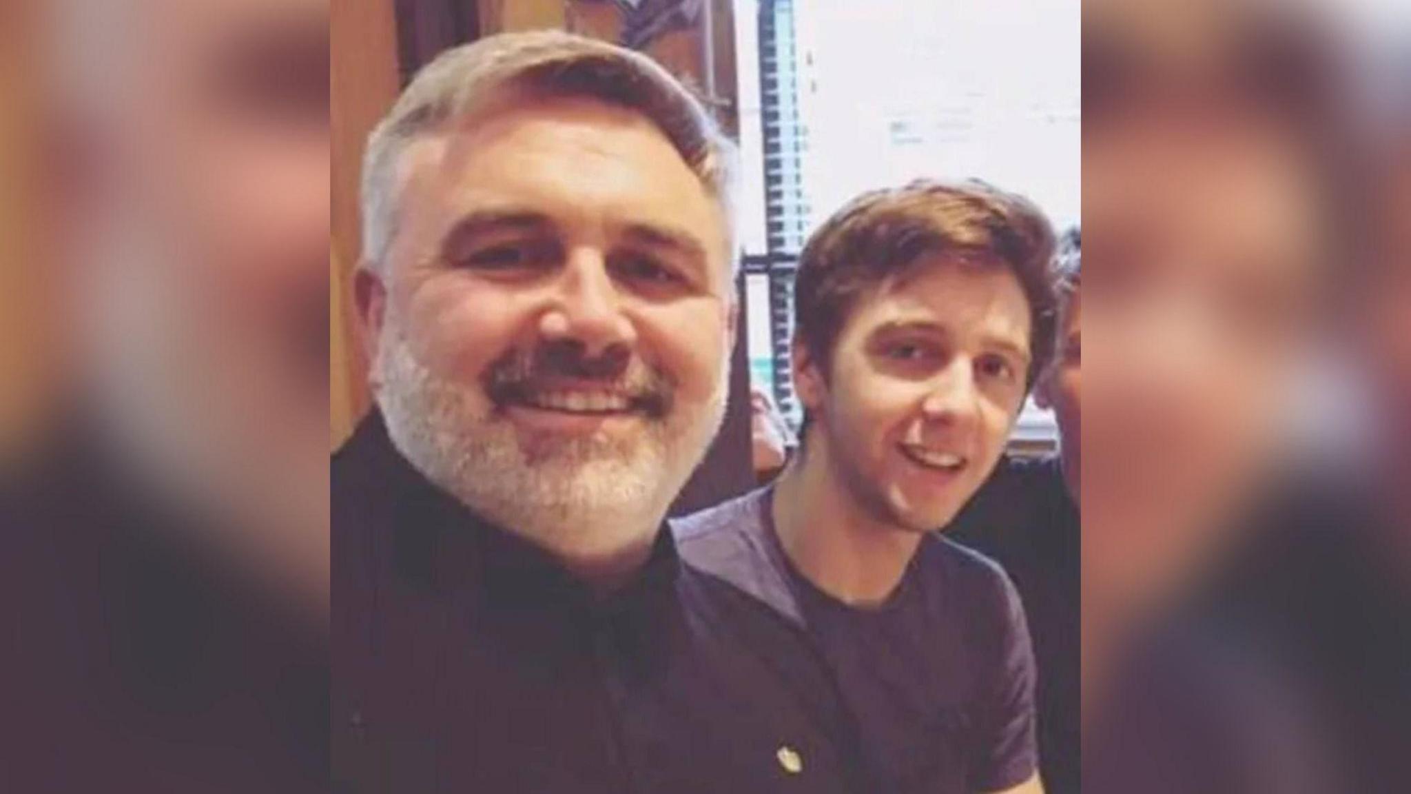 Daniel and Liam look at the camera, both are smiling and wearing dark tops. Daniel has a grey and white beard and short hair and Liam, on the right, has short dark brown hair.