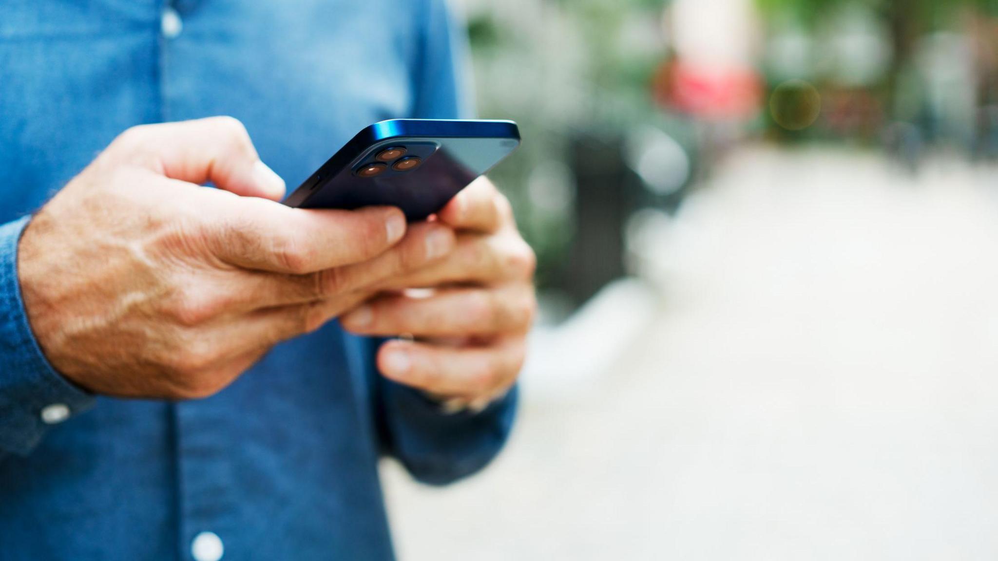 The body of white man pictured as he holds a mobile phone wearing a blue, buttoned-up shirt 