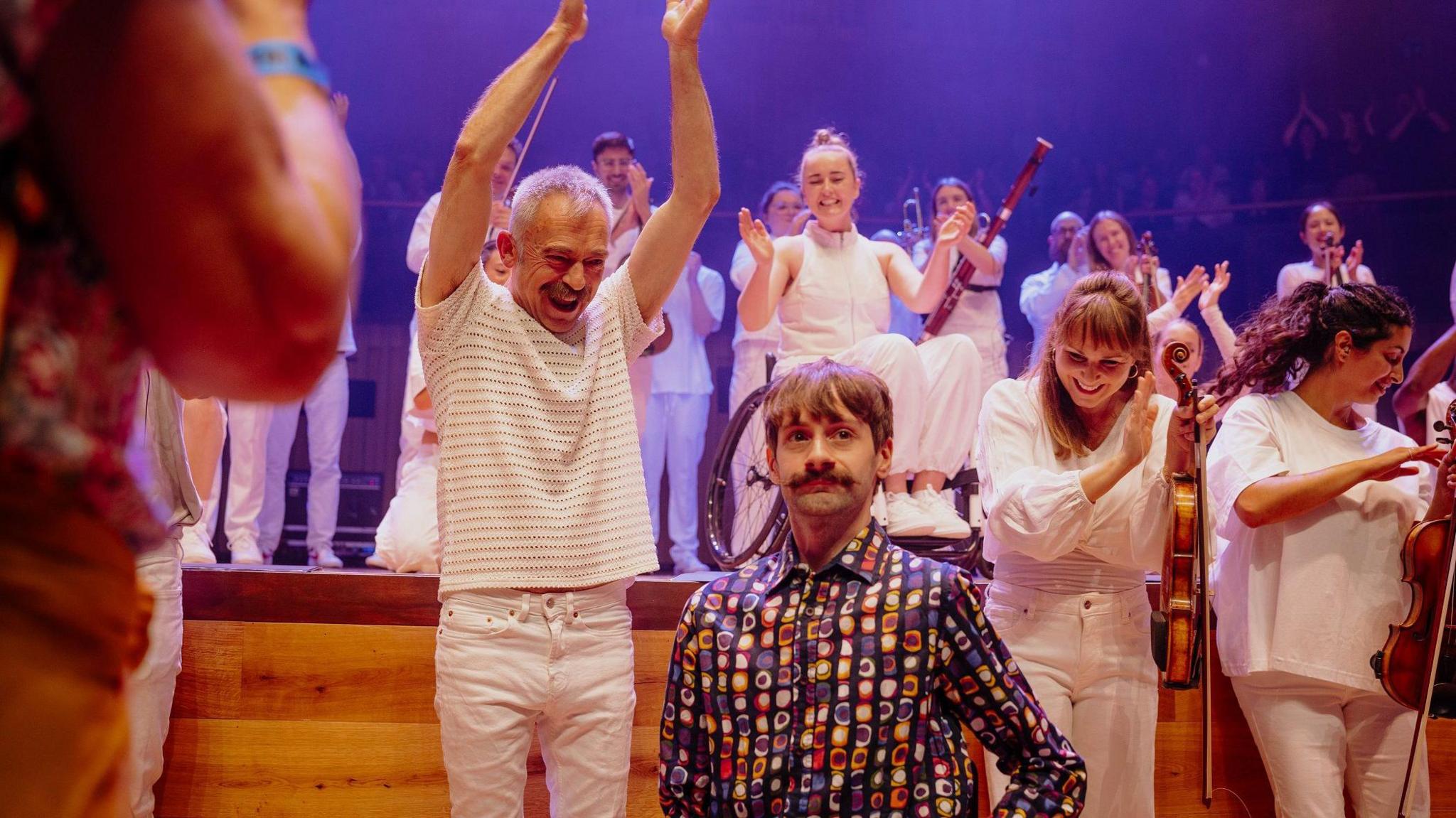 Man in white t-shirt clapping next to another man sitting down in the middle of the photo. They are surrounded by orchestra members holding instruments and all clapping