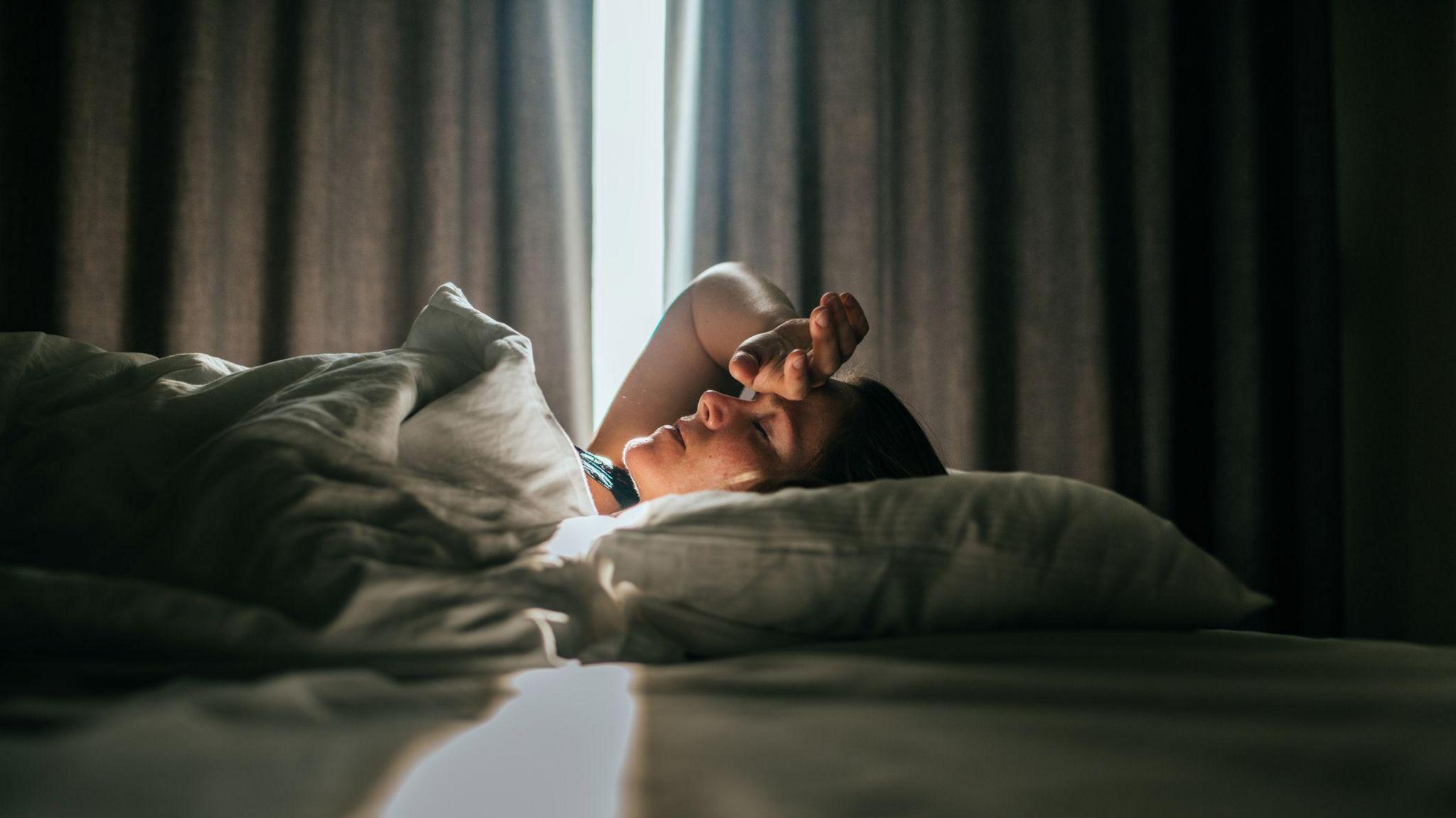 A person lying on their back in bed with their hand on their forehead. They have a pillow under their head and in the background sunlight is coming through grey curtains that are partly opened.