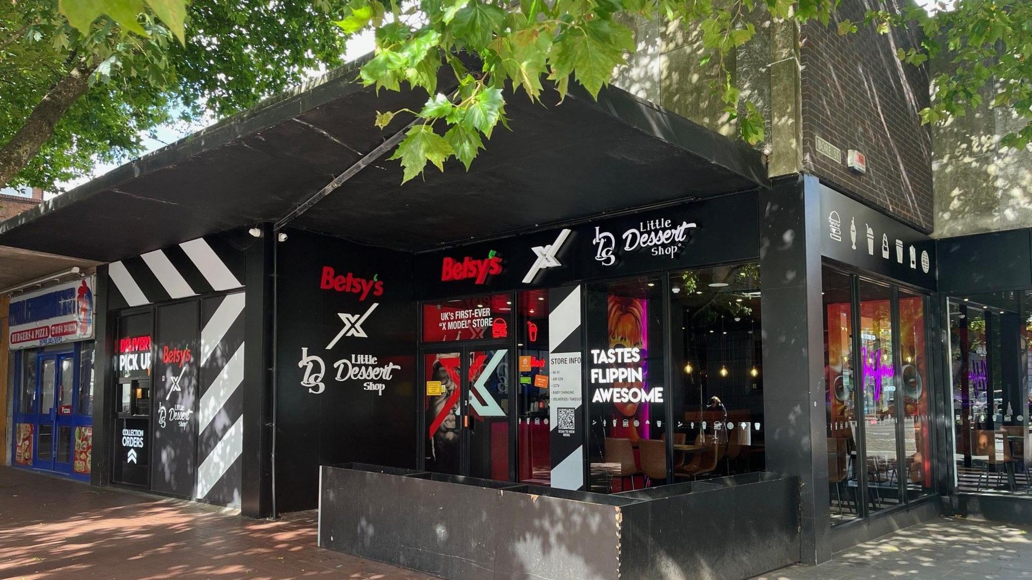 A shop front which has black signage with red and white writing that reads Betsy's X Little Dessert Shop. There are trees in front of the shop.