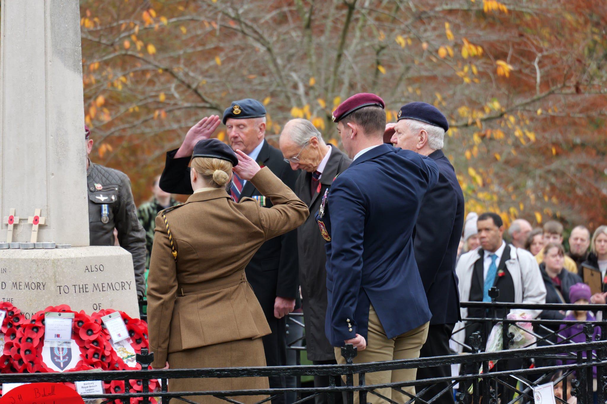 Military personnel and a veteran at the War Memorial in People's Park in Banbury. Crowds are watching the wreaths laying from the sides. It is overcast.