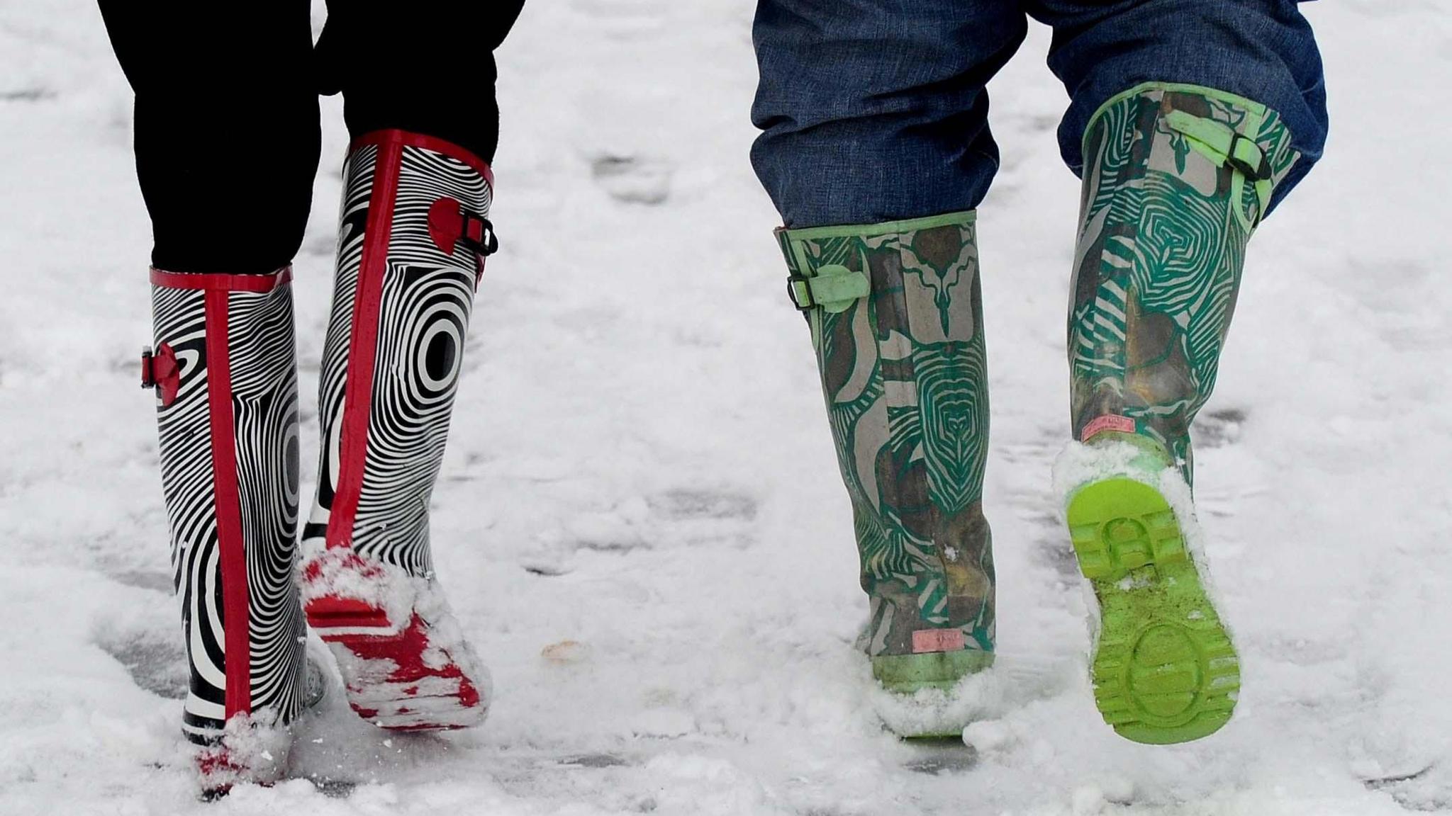 A close up from behind showing the legs of two youngsters walking in brightly-patterned wellies in the snow. The person on the left has black trousers tucked into zebra print and red wellies. The child on the right has boots with a green ad khaki swirling pattern.