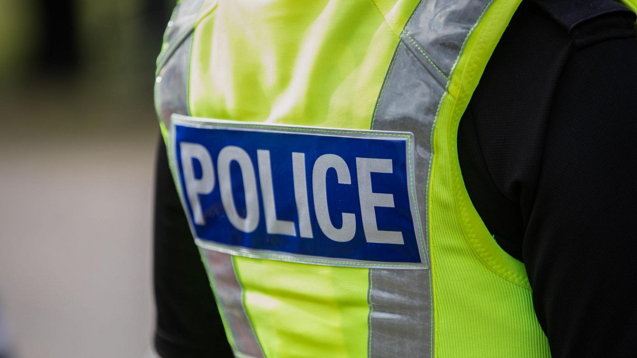 Picture shows the back of a high-vis police jacket. The jacket is mostly bright yellow, with once vertical silver stripe and one blue and silver checked horizontal stripe. The word 'police' is also on the back of the jacket in silver, on a square blue background. 