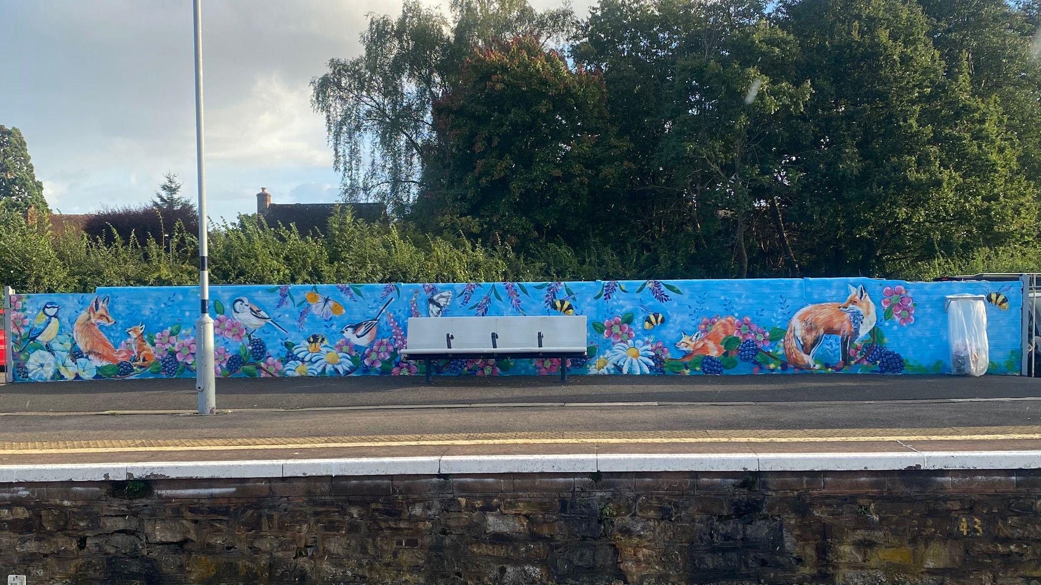 A wide shot of the mural which is painted on a long wall alongside the railway platform. A bench is in the middle of the wall, which is painted blue, with orange and white foxes, colourful birds, and flowers. 