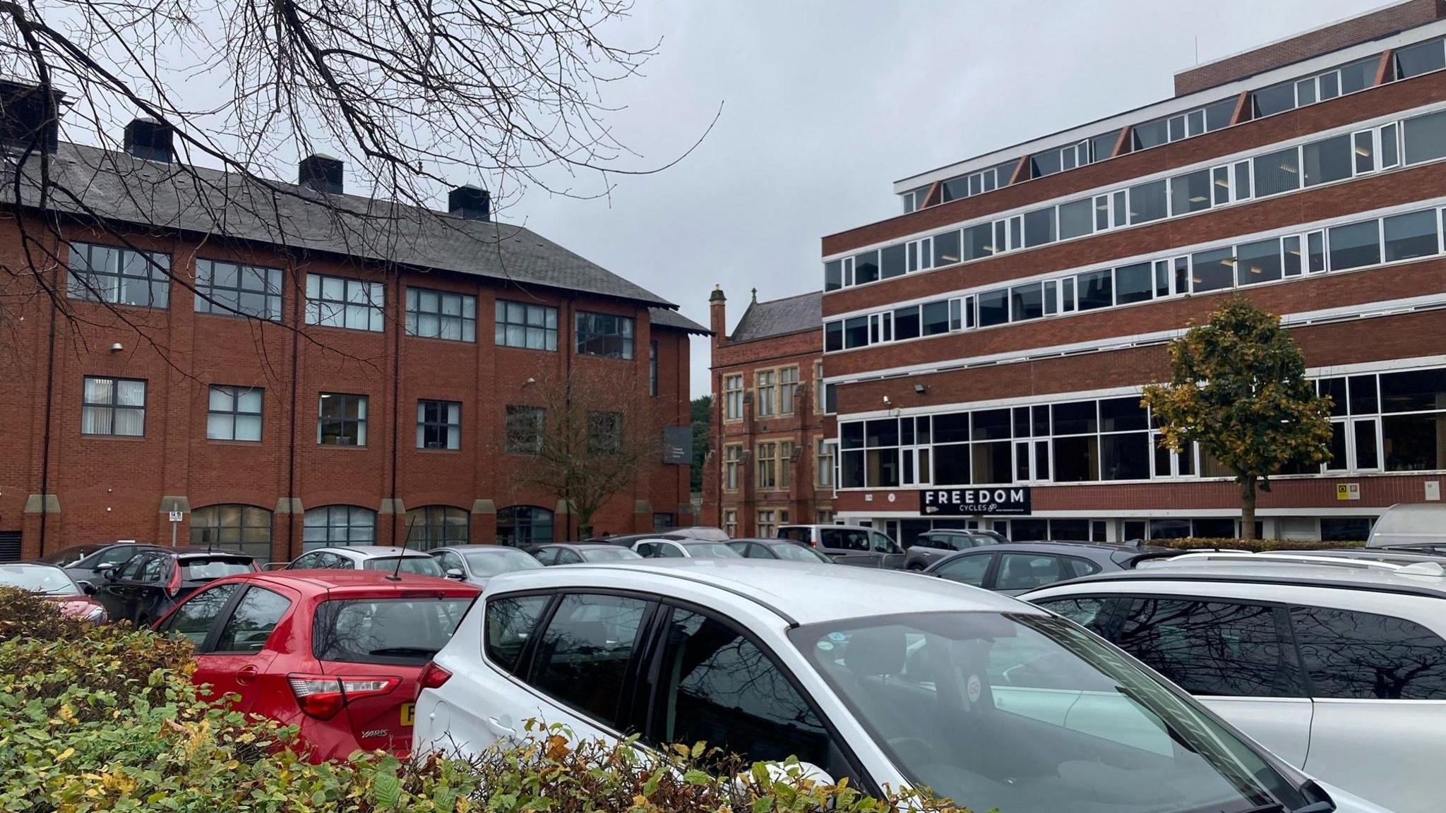 A number of cars parked in a car park. The car park is surrounded by large red-brick buildings. 