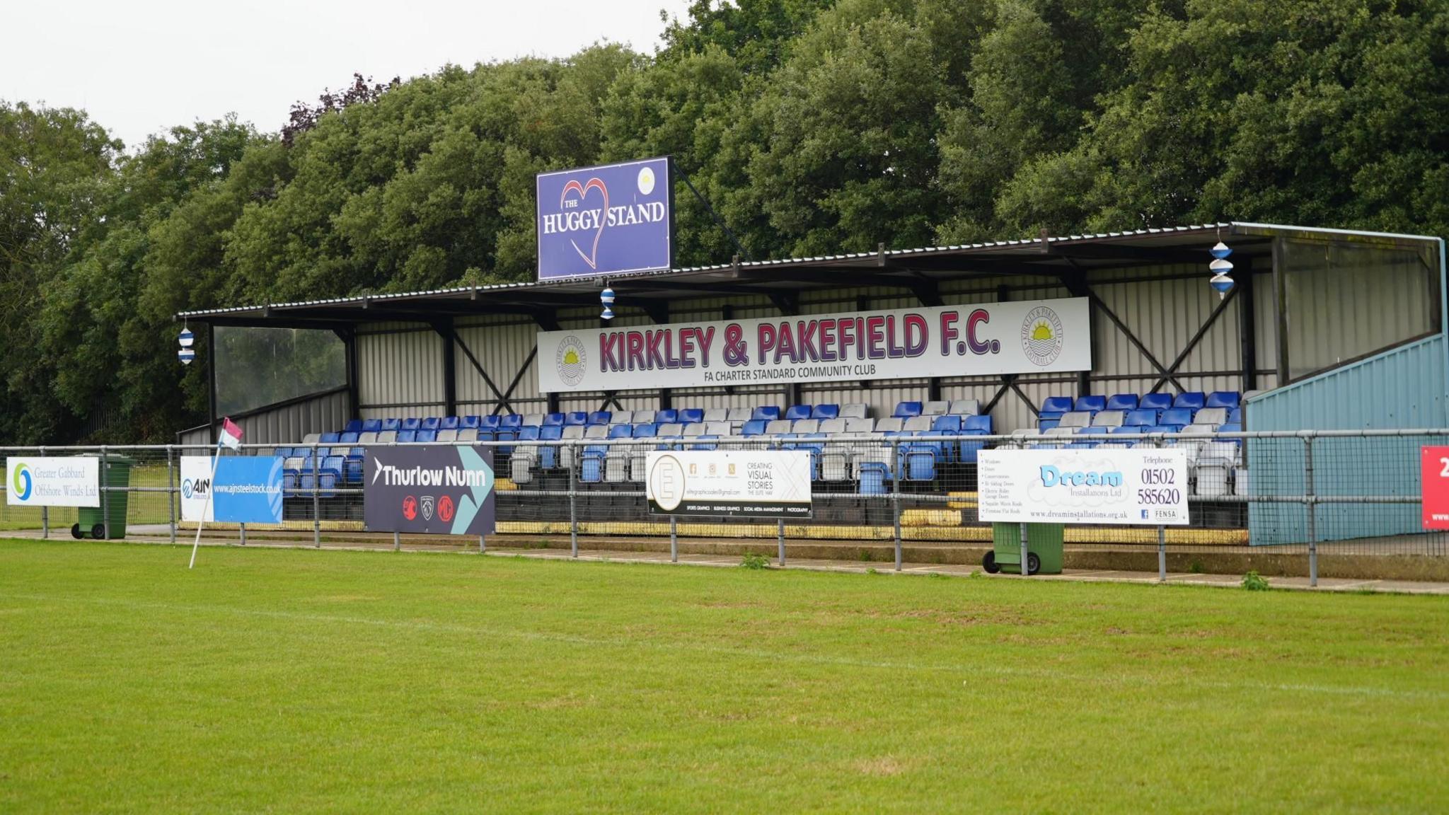 Kirkely and Pakefield Football Club "Huggy" Stand, named after James Hutchinson. It has a row of seats five deep in blue and white colours and there are various advertising boards attached to a metal framed fence in front of the corrugated iron stand. You can see the green pitch in front of it.