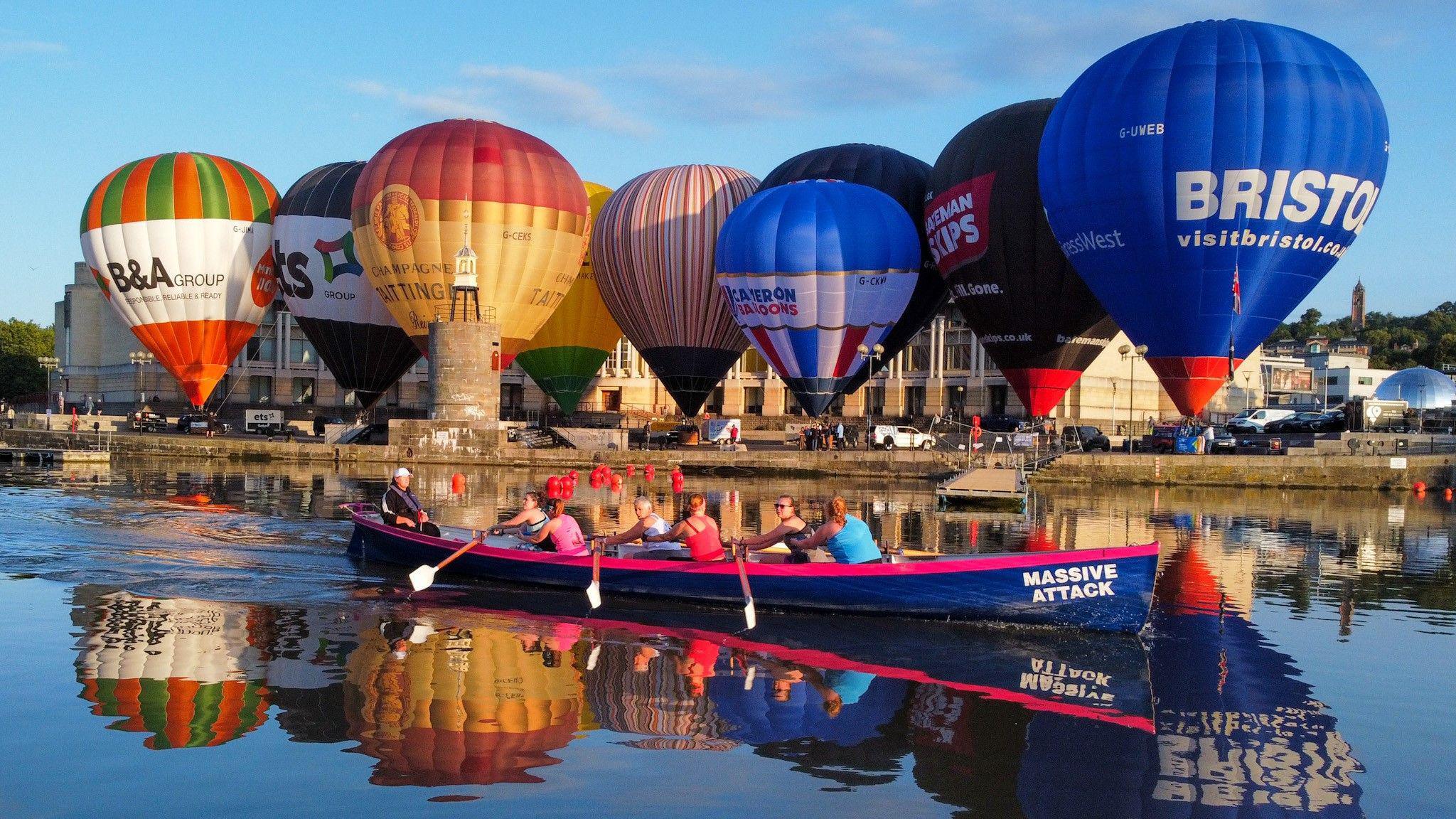 Several hot air balloons tethered to the ground in Bristol Harbour. There is a small row boat in the foreground of the picture.