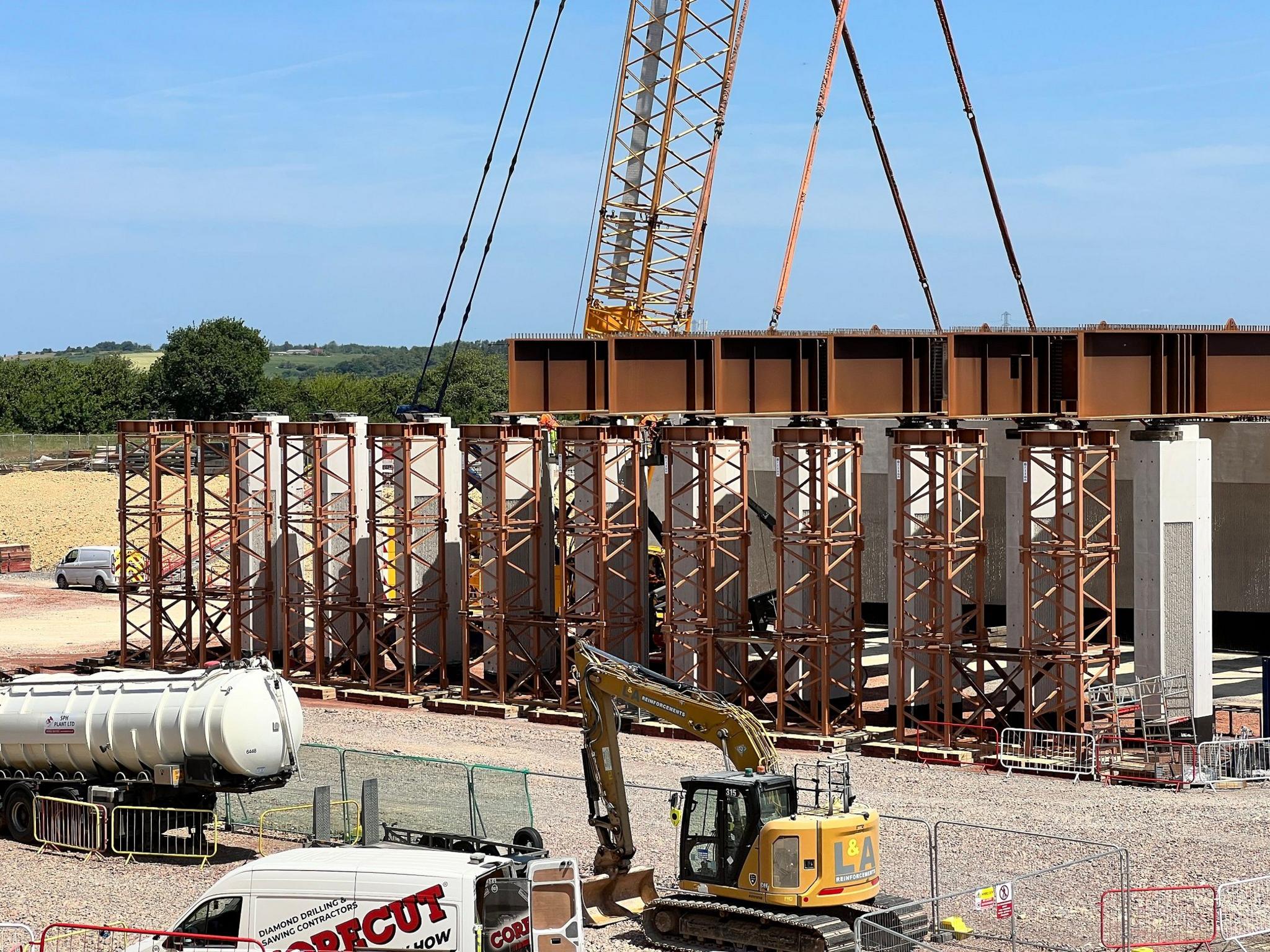 A large bridge structure made of brown steel beams on a building site. It has not yet been lifted into place but is surrounded by construction vehicles including vans, a tanker and a yellow digger.