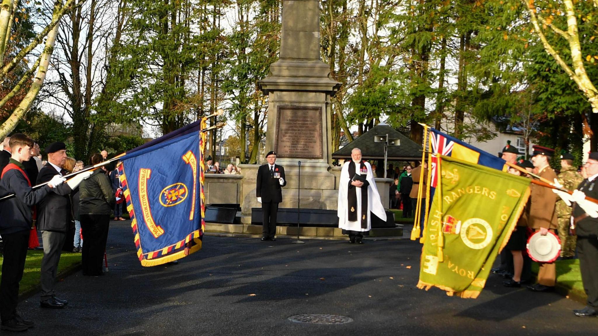 Flags are being held in front of a war memorial. A man standing in front of the monument is wearing a poppy and a minister is standing next to him, holding a book. Some people are wearing army uniforms. 
