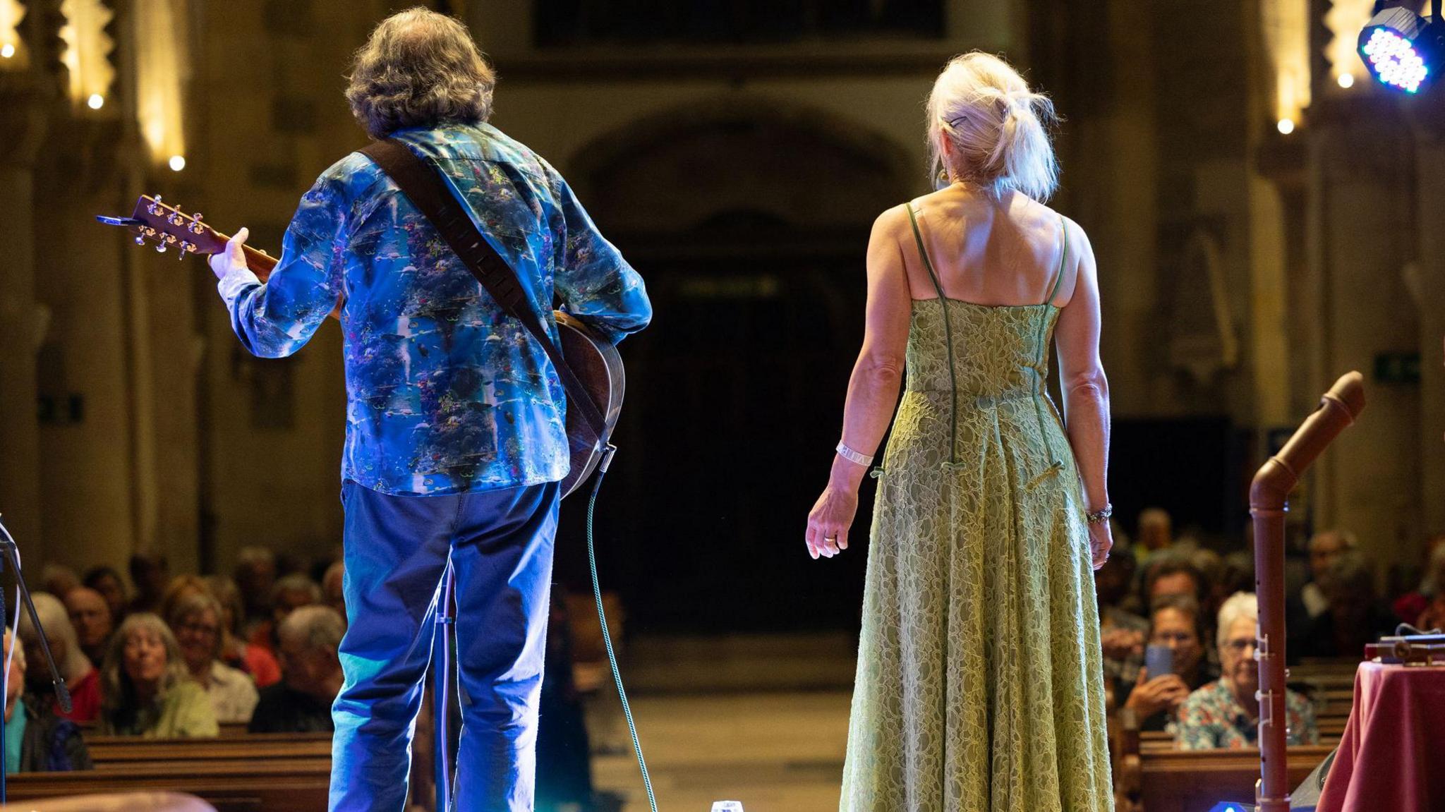 Paul Walker with a guitar and Karen Pfeiffer singing, stood at the front of a church 