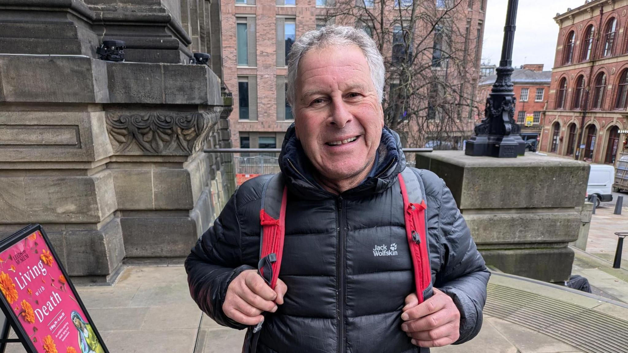 A middle-aged man with a black coat and rucksack, with red straps over both shoulders, smiles into the camera.