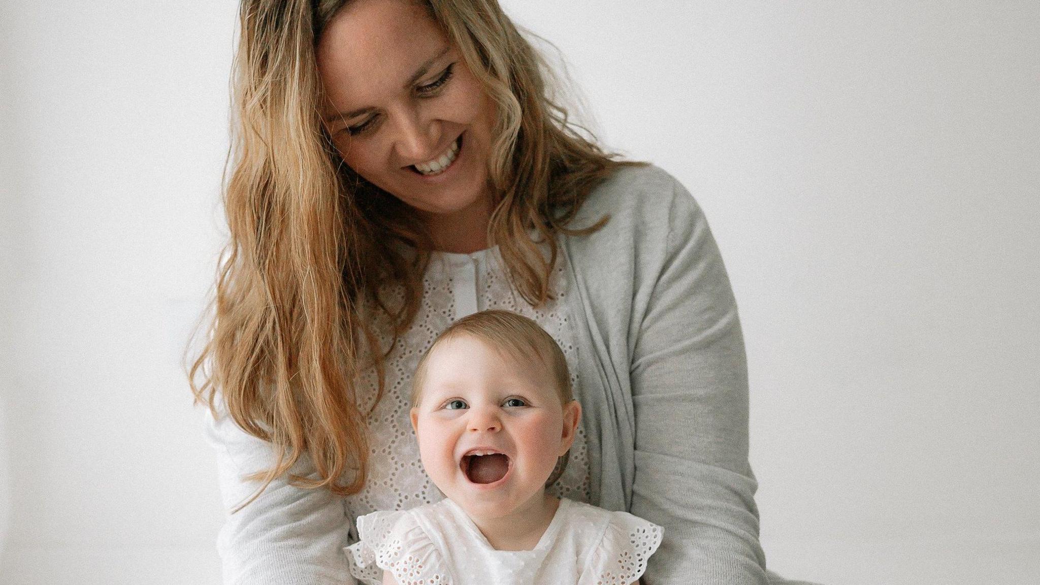 A woman with long blonde hair looks down at a smiling child on her lap. They are both dressed in white and sitting against a white background.