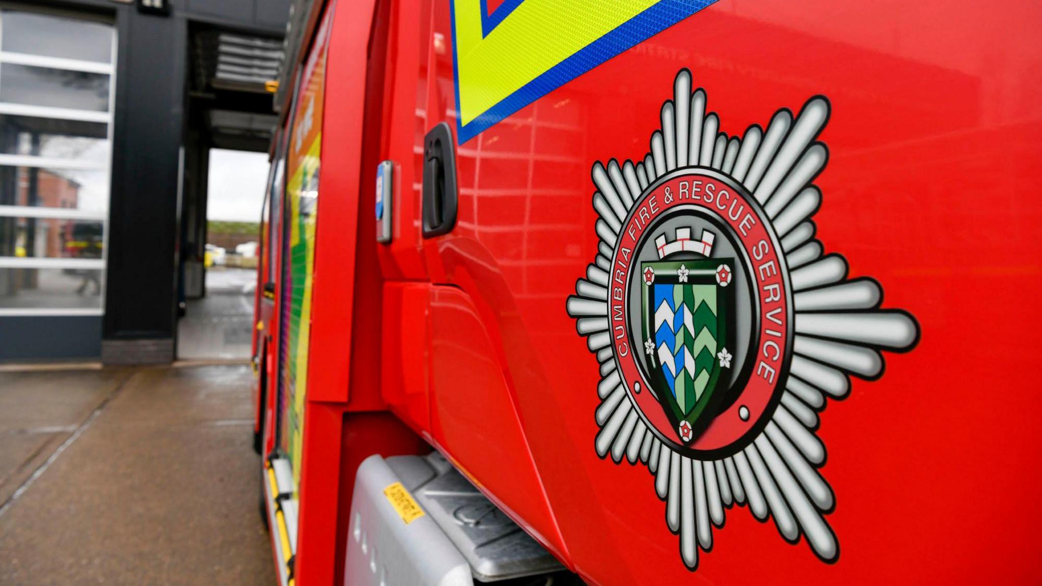 The side of a red fire engine, with the emblem on that reads 'Cumbria Fire & Rescue Service'. The engine also has yellow and blue stripes and is parked inside a fire station.