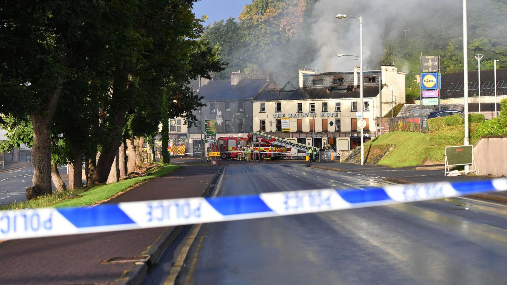 Police tape in the forefront of the image. In the background is the derelict Railway Hotel in Enniskillen. There are firefighters and a fire engine outside. There is smoke coming out of the top of the building and fire damage is visible on the roof. 