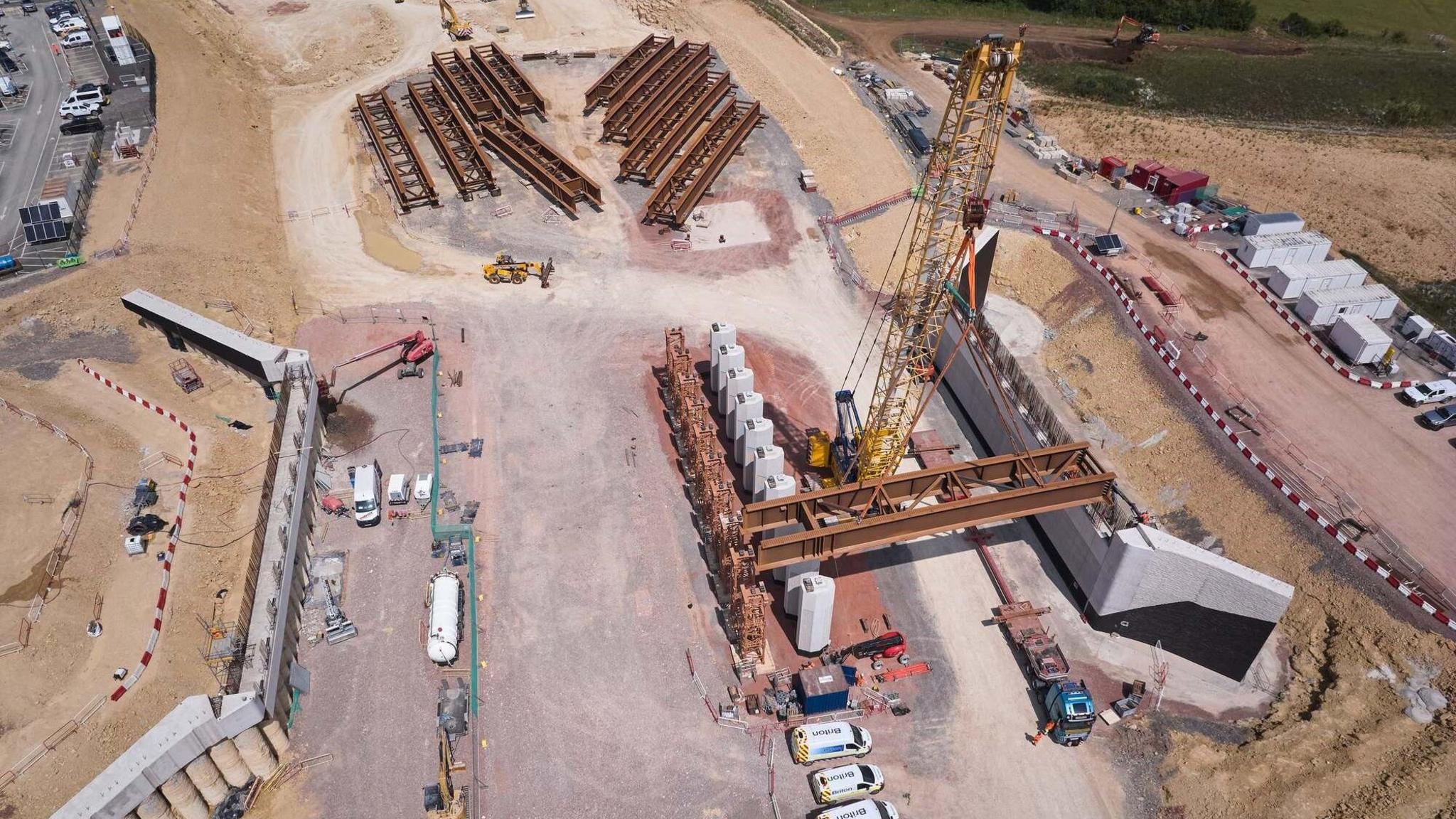 An aerial shot of the area around the bridge construction, with a large yellow crane and several massive steel beams which dwarf the cars and vans parked around the site. 