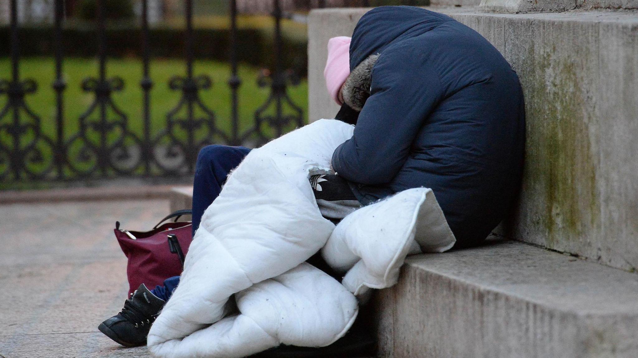 A faceless person is sprawled out over a step wearing a thick winter coat and carrying a sleeping bag