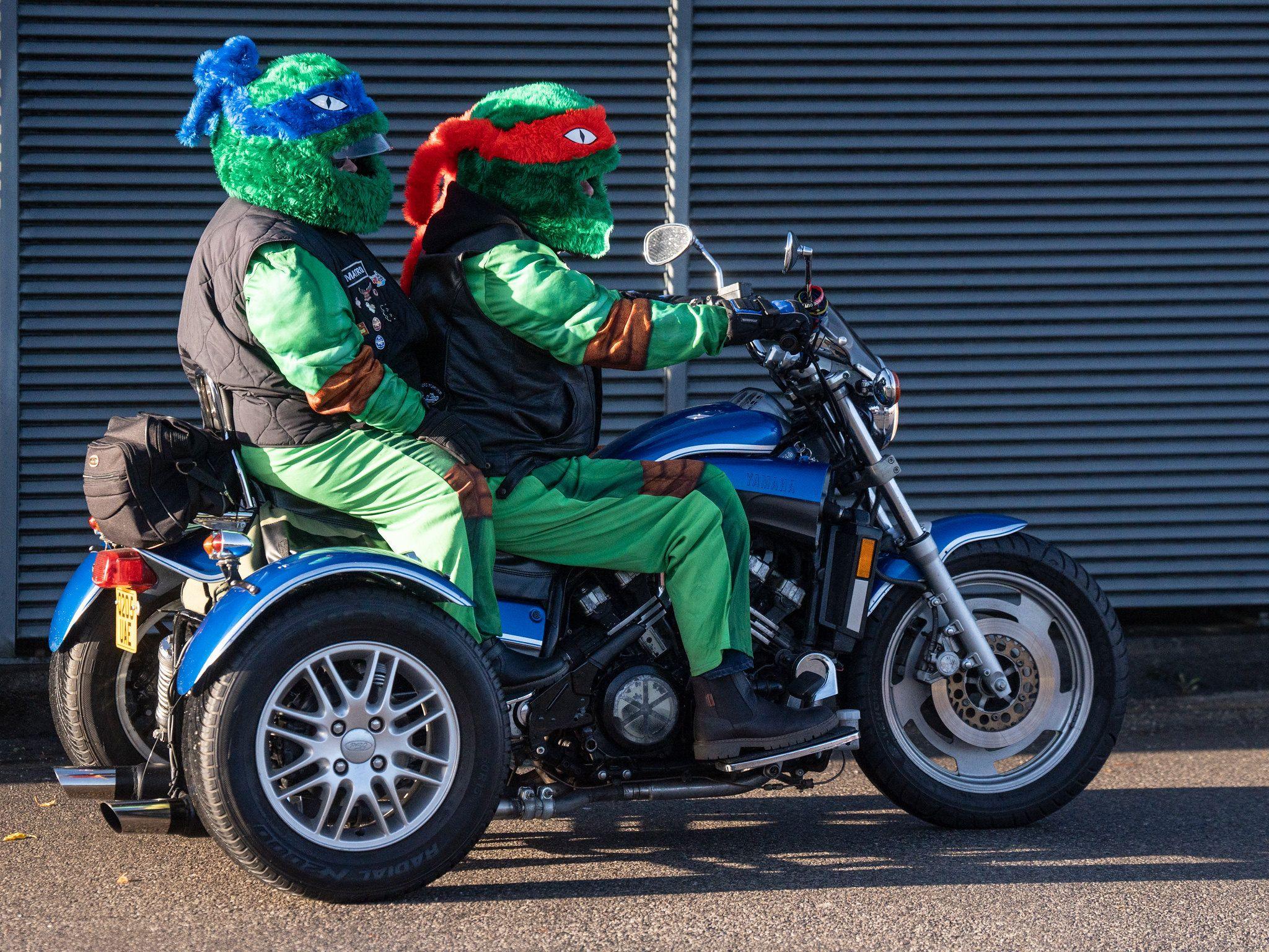 Two people dressed as Teenage Mutant Ninja Turtles are sat on a blue three-wheeled bike.