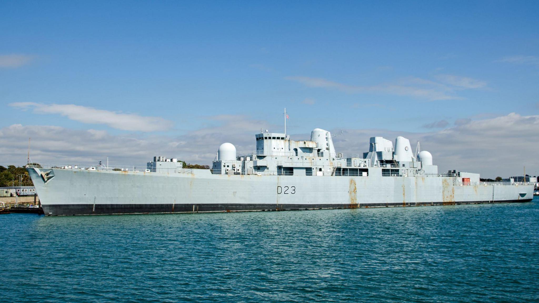 An enormous grey warship docked on a quayside on a sunny day. The hull has a couple of rust marks.