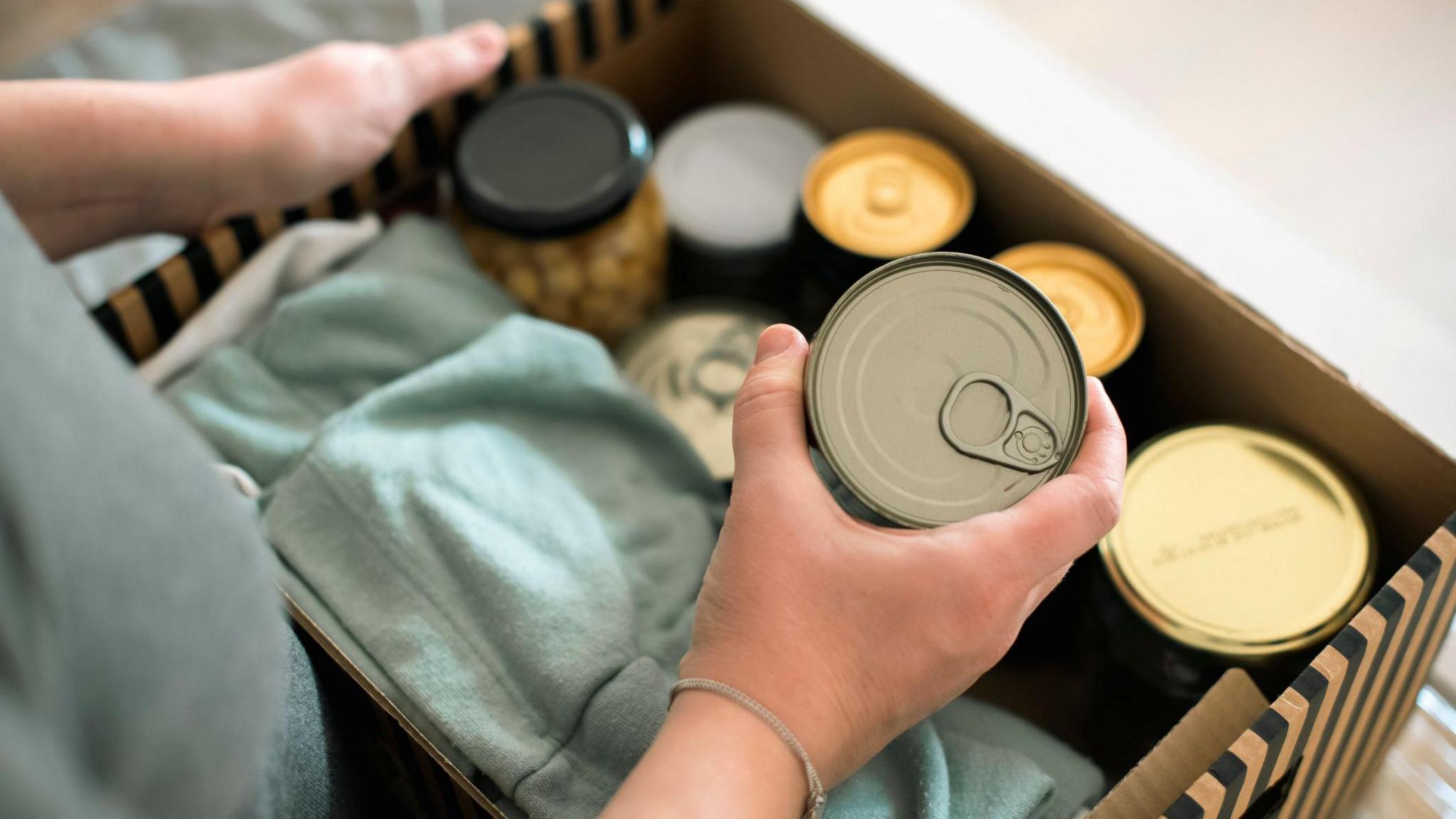 Hands holding a canned food item above an open cardboard box filled with other canned items. The box has a striped design along its edges.