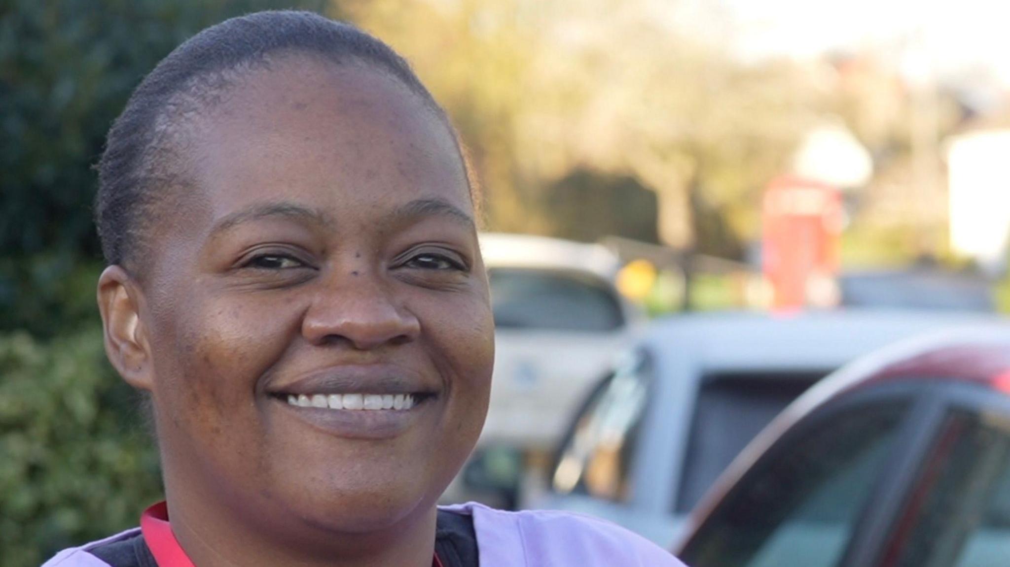 Dephine Nhidza stands on the pavement with cars parked beside and behind her. She has dark hair that is tied back and is smiling at the camera. She is wearing a light purple top.