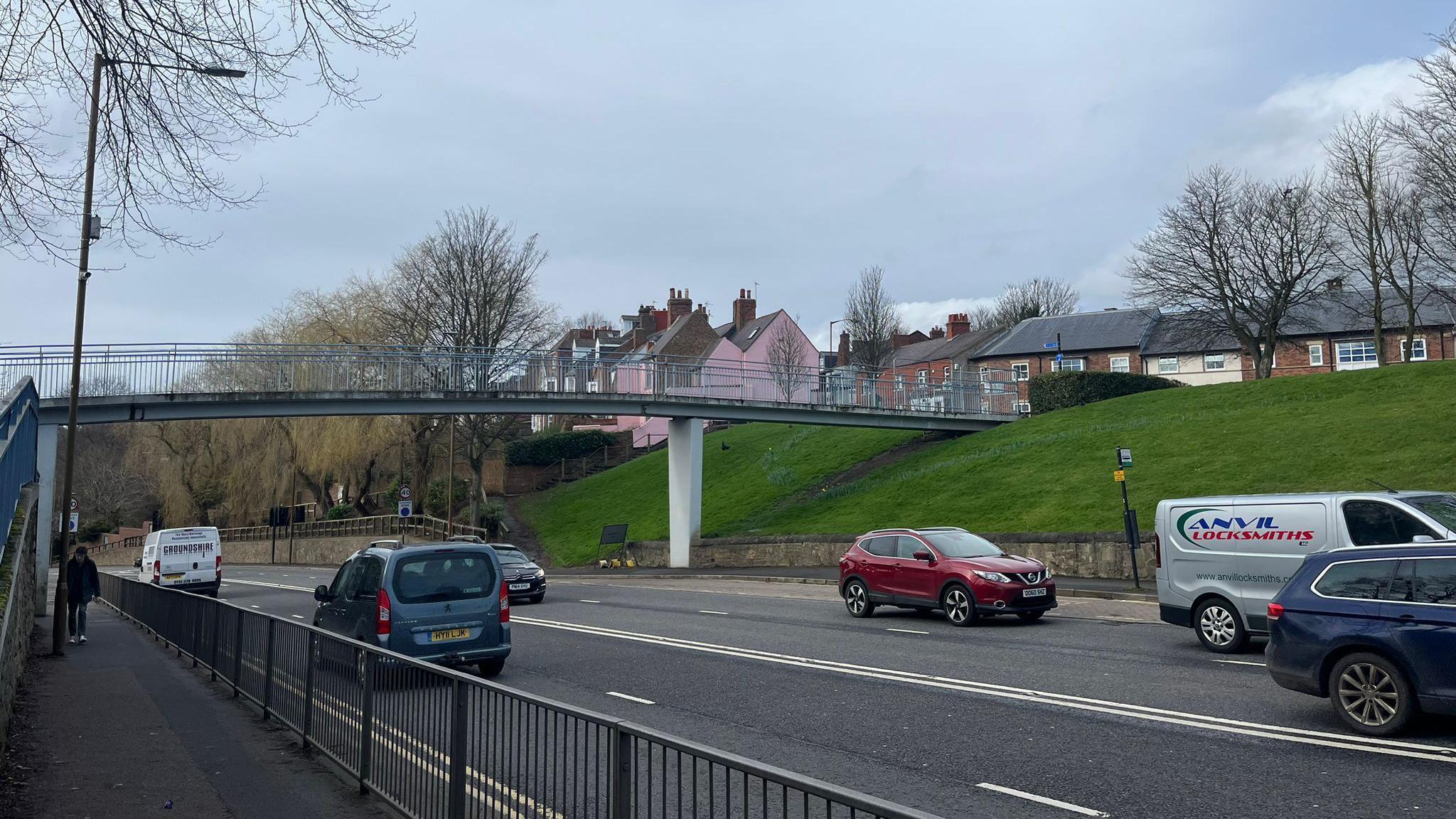 A grey bridge with blue railings suspended over a busy road with traffic moving in both directions. A grassy bank and houses can be seen to the right.